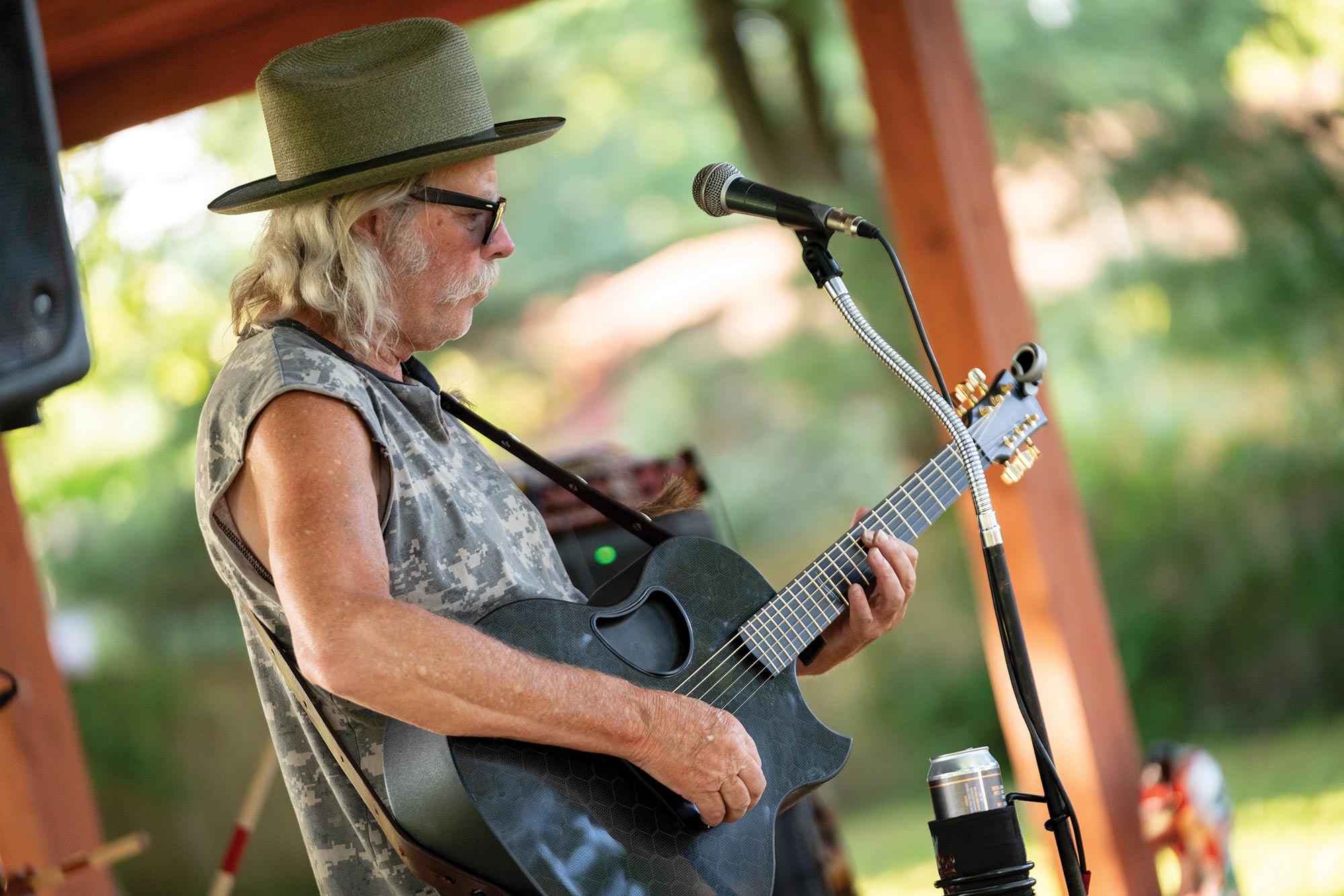 A person with long grey hair, wearing a hat and sunglasses, playing an acoustic guitar and standing in front of a microphone outdoors.