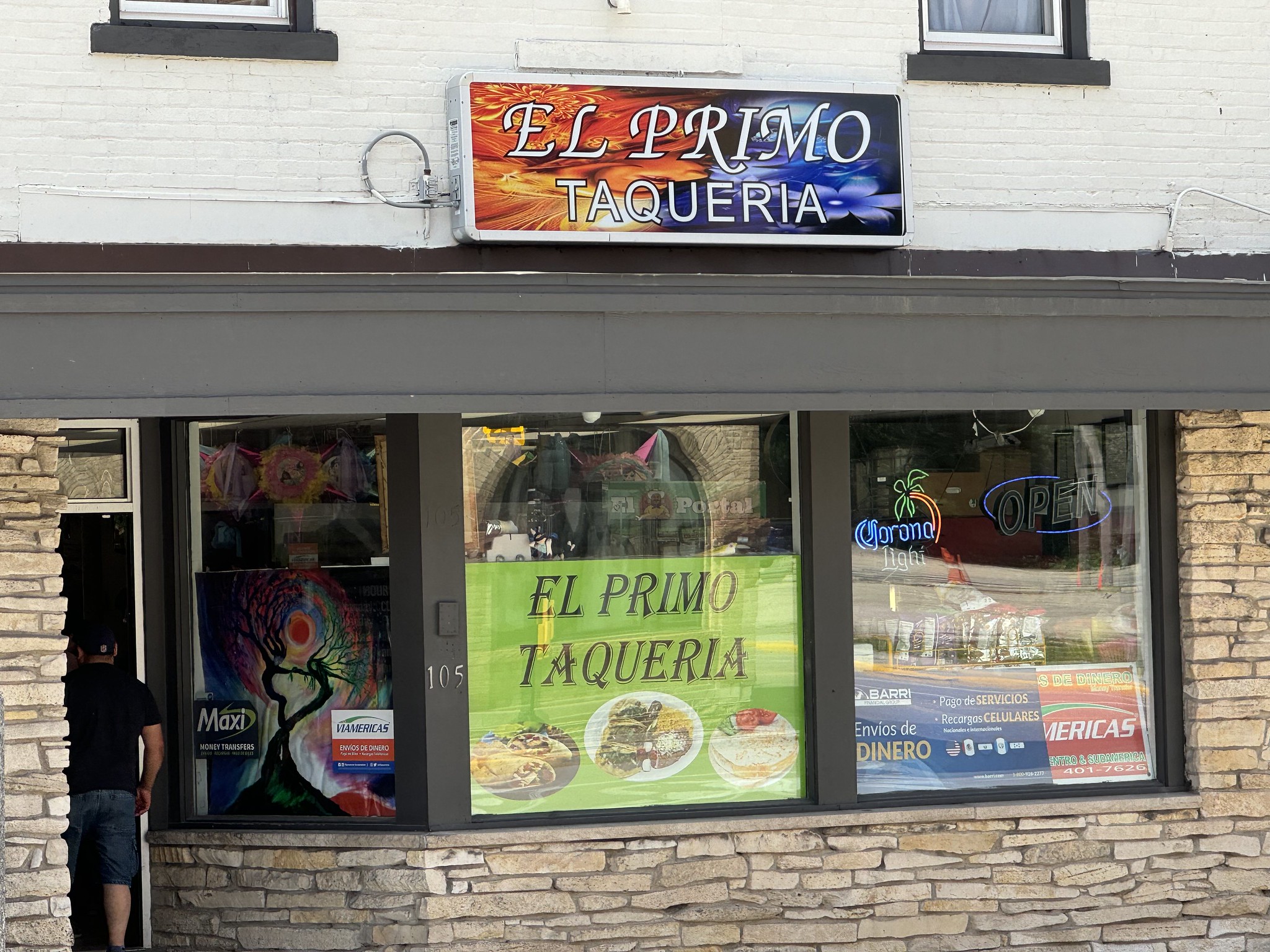 Front view of "El Primo Taqueria" showing the exterior with signage, a banner advertising food items, and a person entering the doorway. The windows display various signs and promotional materials.