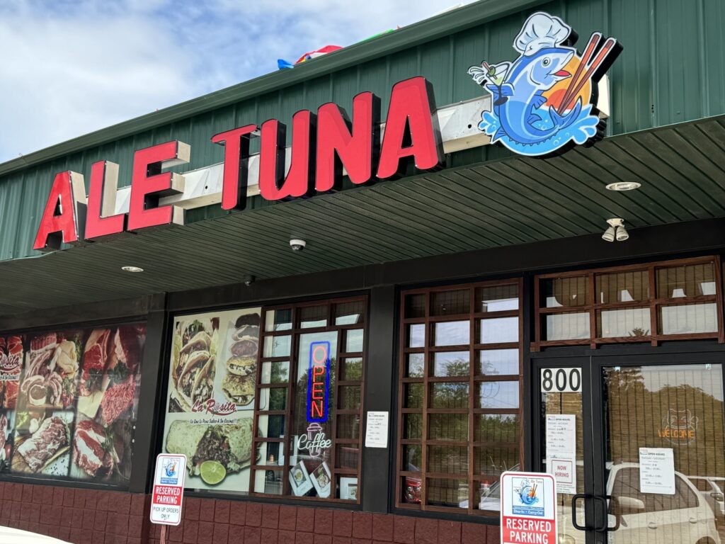 Facade of a restaurant named "Ale Tuna" with a fish logo. The storefront features large windows displaying food images, a lit "Open" sign, and reserved parking for takeout in front.