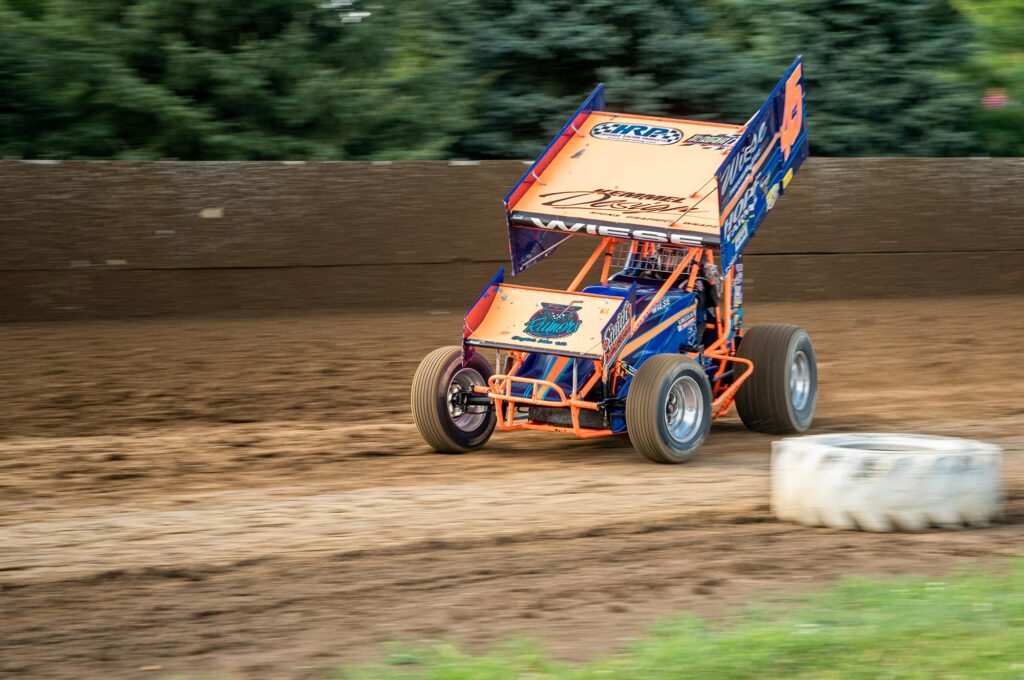 A sprint car with large wing spoiler races on a dirt track, surrounded by a brown wall and greenery in the background.