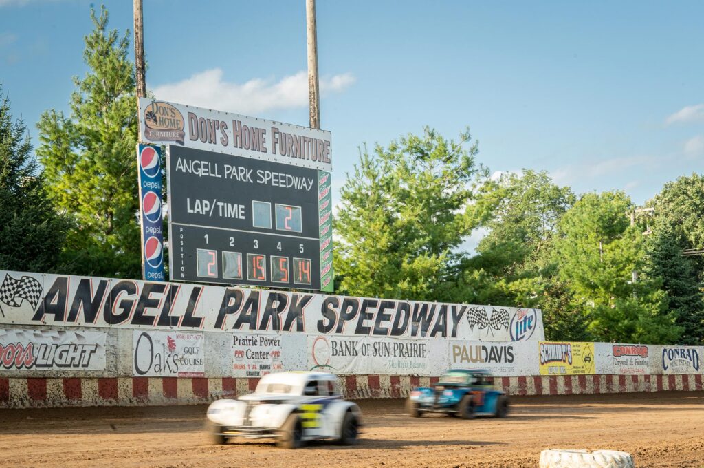 Race cars on a dirt track at Angell Park Speedway with a scoreboard showing lap times and sponsorship banners in the background.