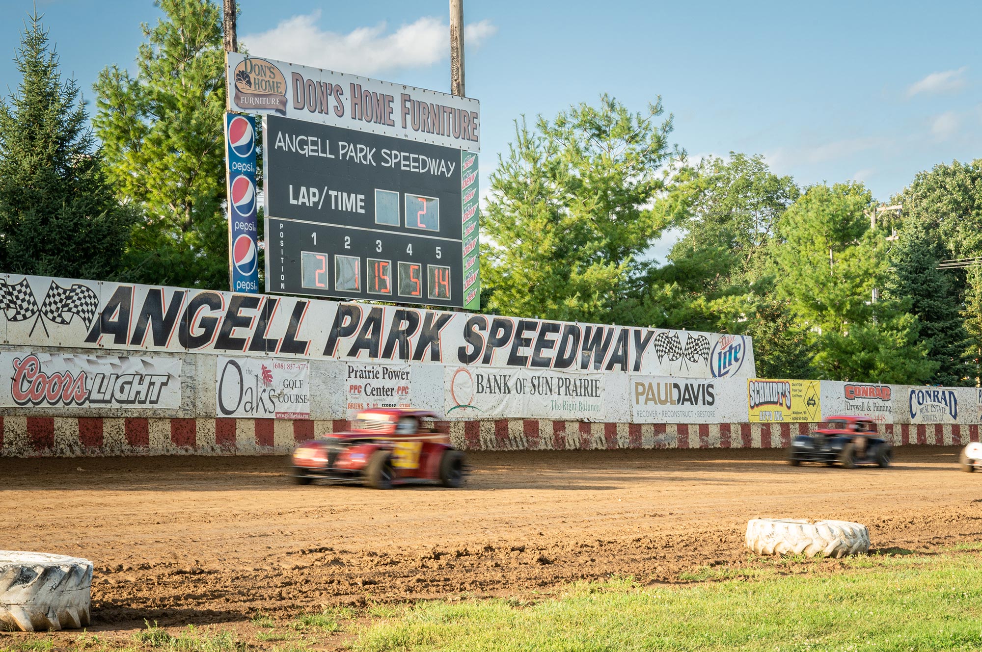 Three race cars speed on a dirt track at Angell Park Speedway. The scoreboard displays lap times and sponsors’ advertisements line the track. Trees and a clear sky are visible in the background.