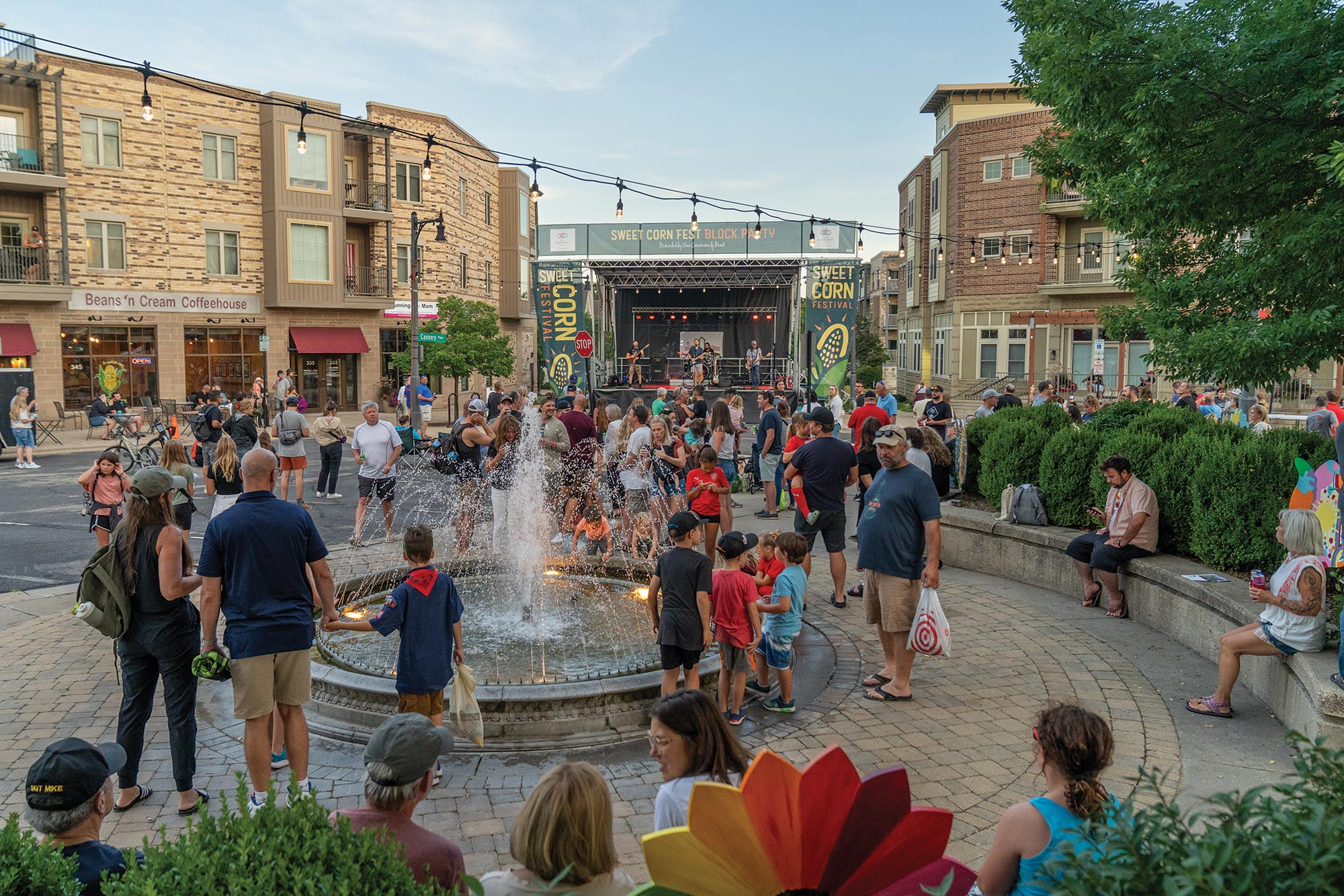 A diverse crowd gathers in a lively outdoor plaza with a fountain, enjoying a live music performance on stage. People are talking, walking, and relaxing on benches under strings of lights.