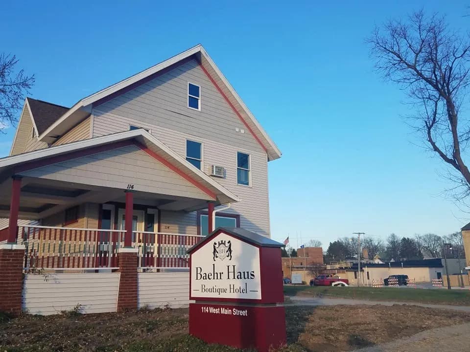 A two-story house with a sign reading "Baehr Haus Boutique Hotel, 114 West Main Street" in front. The house has a porch and is surrounded by a lawn and trees.