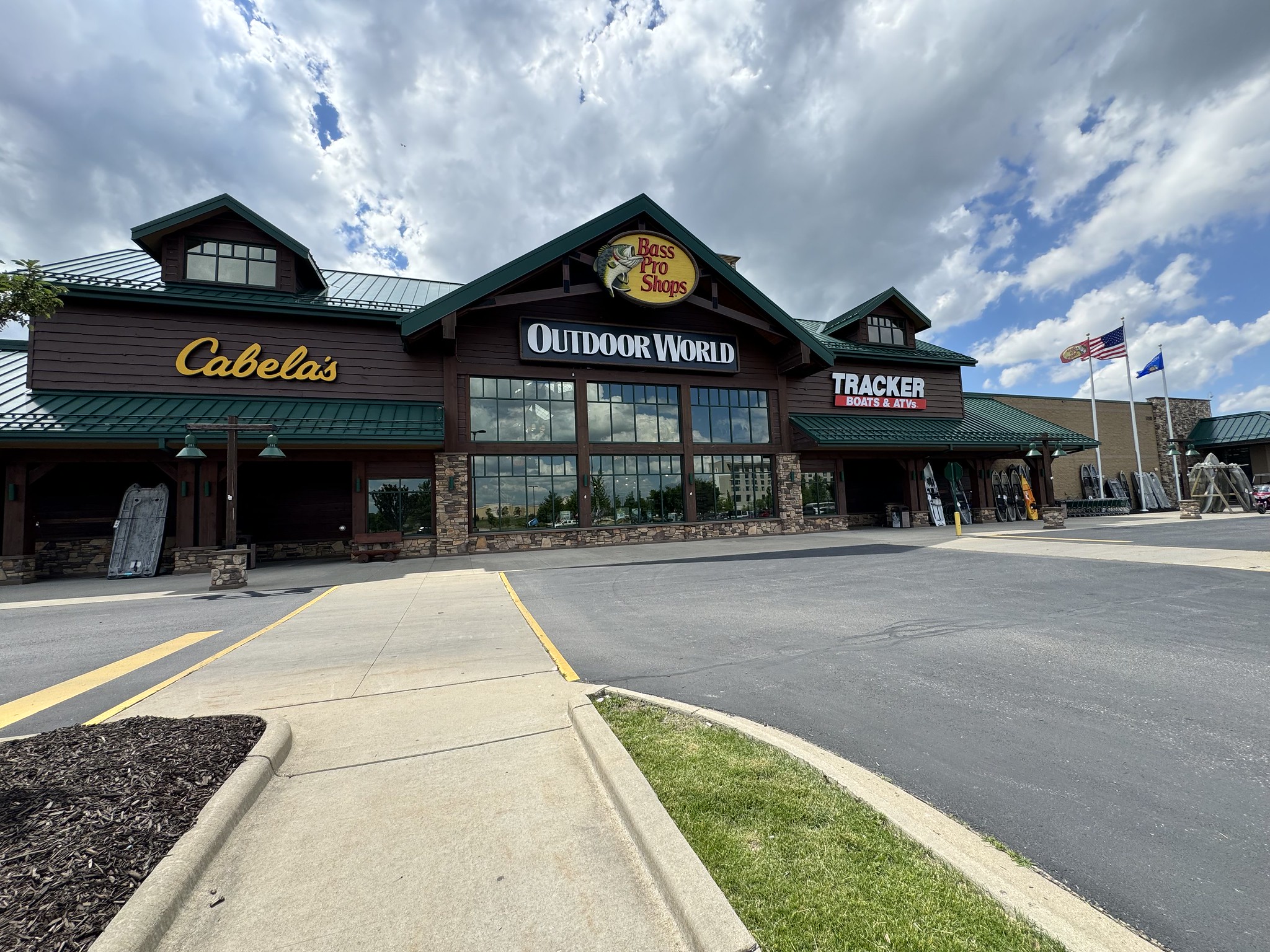 A large retail store with signs for Cabela's, Bass Pro Shops, Outdoor World, and Tracker Boats & ATVs. The building has a green roof, stone accents, and multiple American flags. The sky is partly cloudy.