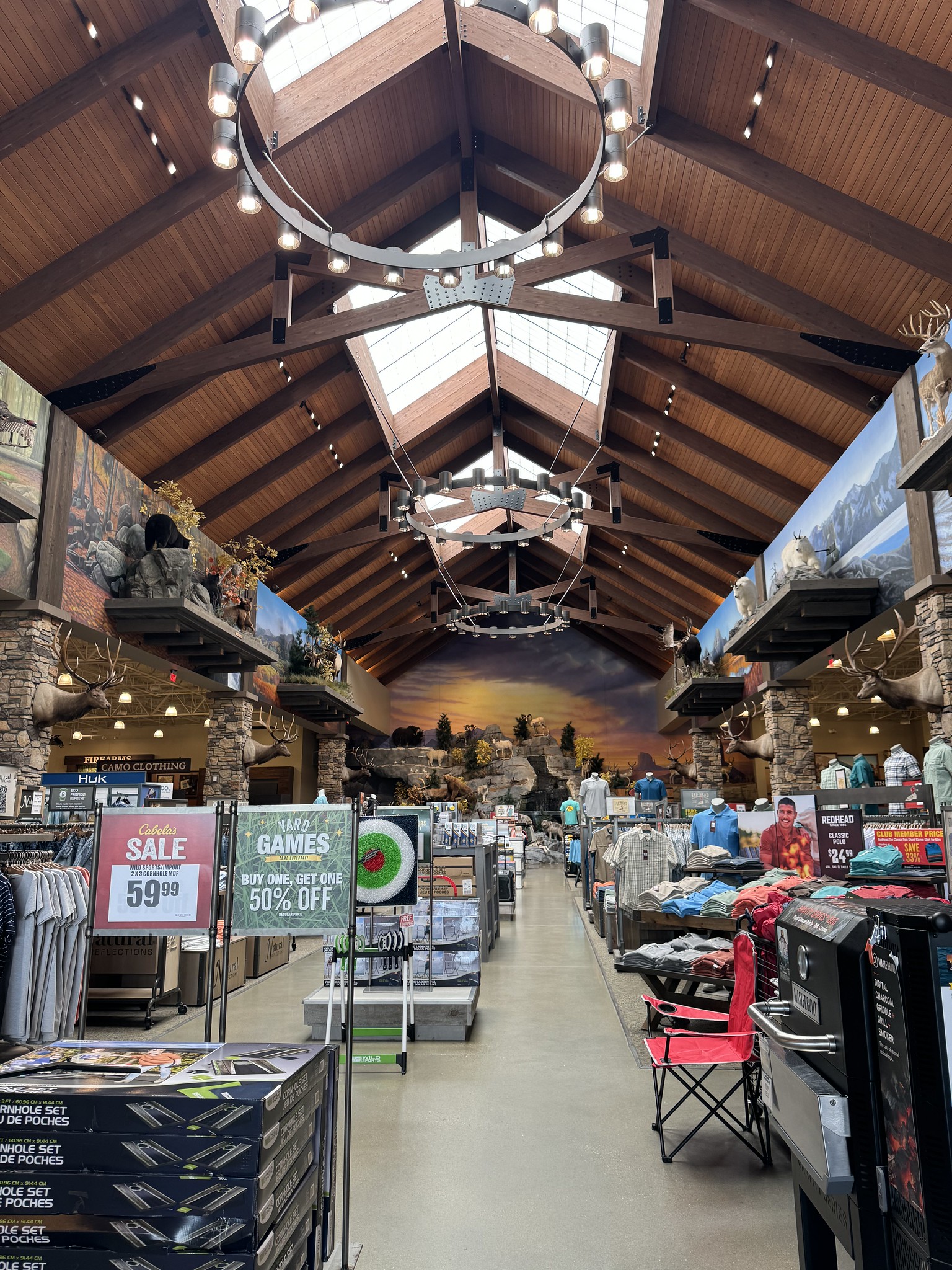 An outdoor-themed store interior features high wooden ceilings, chandeliers, clothing racks, sale signs, and camping gear. A nature mural spans the back wall.