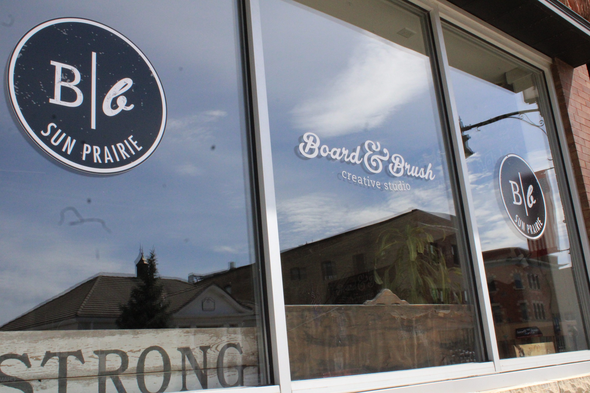 Exterior view of a storefront with large windows displaying the logos of "Board & Brush Creative Studio" and "B|b Sun Prairie.