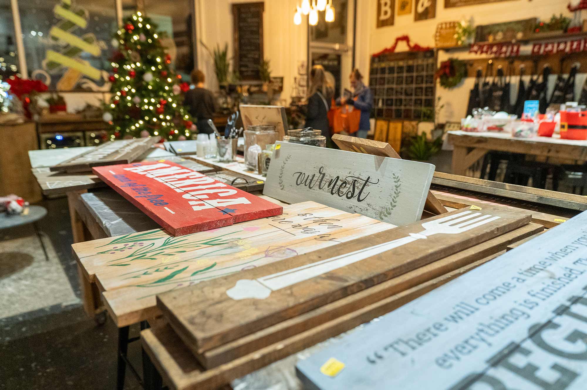 An indoor holiday-themed craft workshop featuring wooden signs and decorations on a table, with a Christmas tree and people in the background.