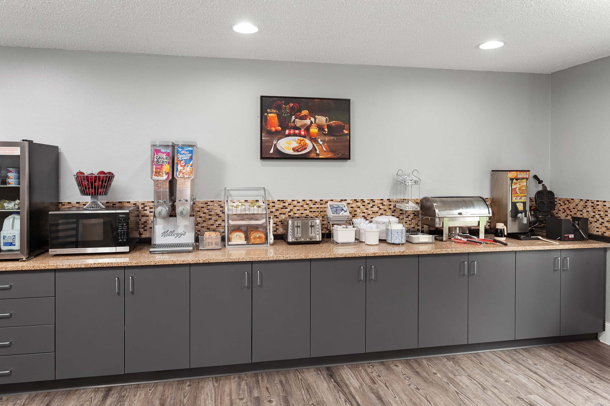 A breakfast bar with various appliances, cereals, a waffle maker, bread, a microwave, and condiments against a wall with cabinets and a framed picture above. Wood flooring is visible.