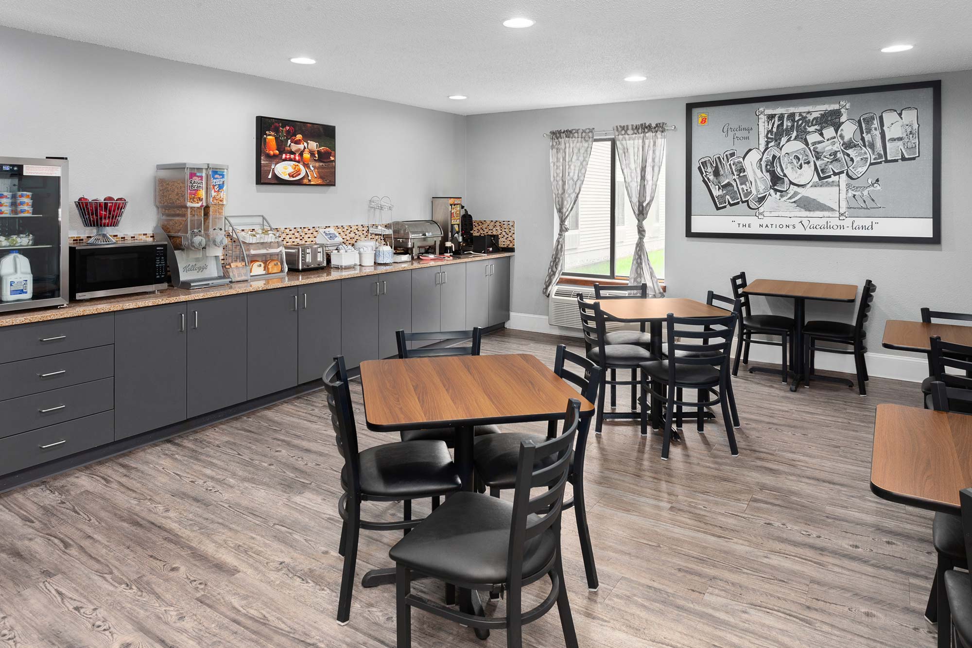 A dining area with wooden tables and chairs, a breakfast bar with cereals, a waffle maker, a microwave, and a coffee machine. The room has a painting and a large window with curtains.