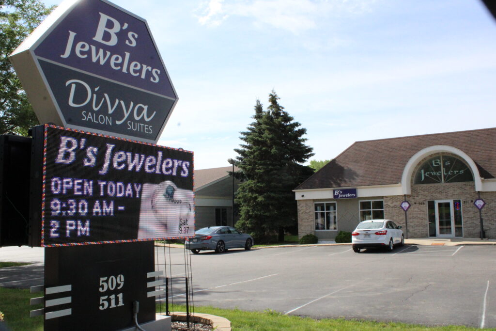 Image of B's Jewelers and Divya Salon Suites. A sign in front reads "B's Jewelers, OPEN TODAY 9:30 AM - 2 PM." The building is in the background with a parking lot in the foreground.