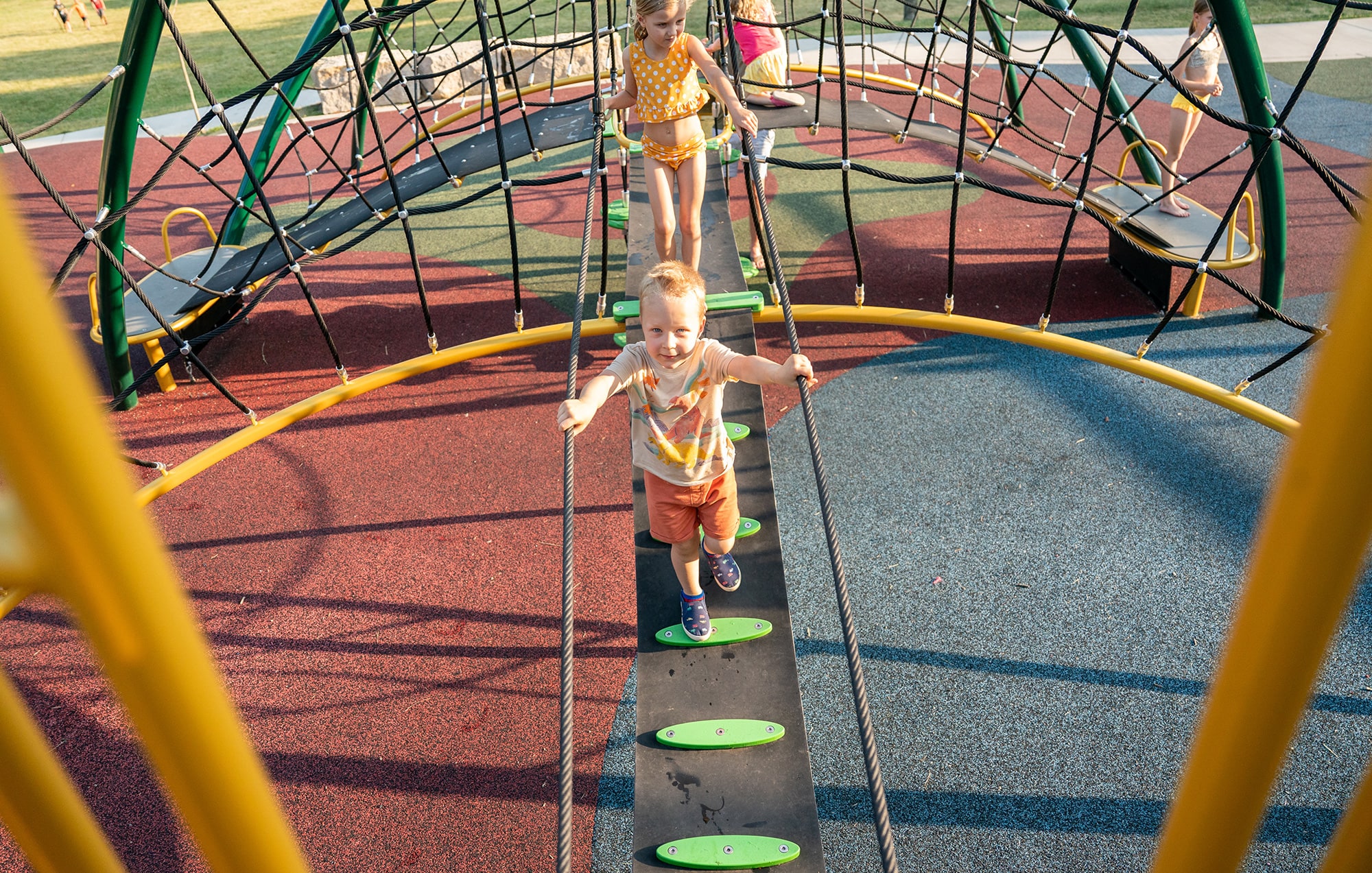 Children climb and play on a colorful, rope-based playground structure on a sunny day.