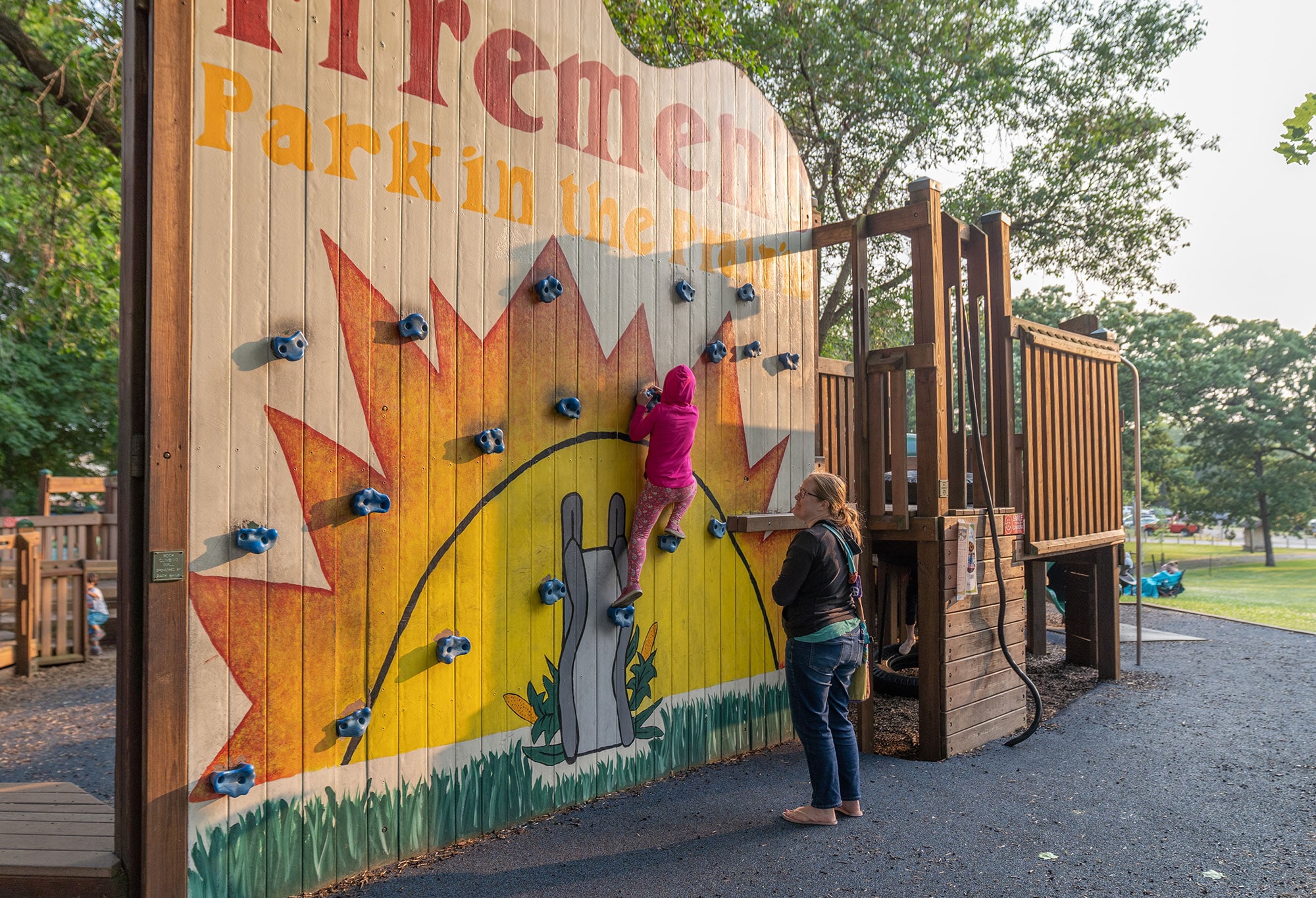 A child climbs a wooden wall with handholds at Fireman's Park while an adult stands nearby watching. Trees and playground equipment are visible in the background.