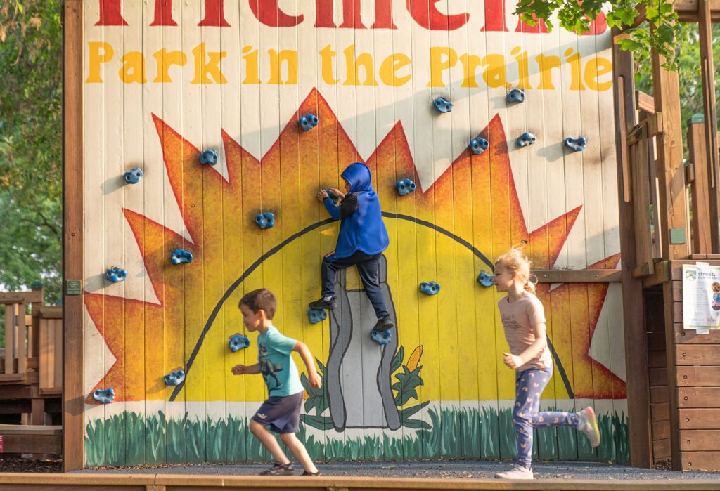 Three children play on a climbing wall and running track at a park with a sign reading "Park in the Prairie" in the background. One child climbs, while the other two run past the climbing wall.