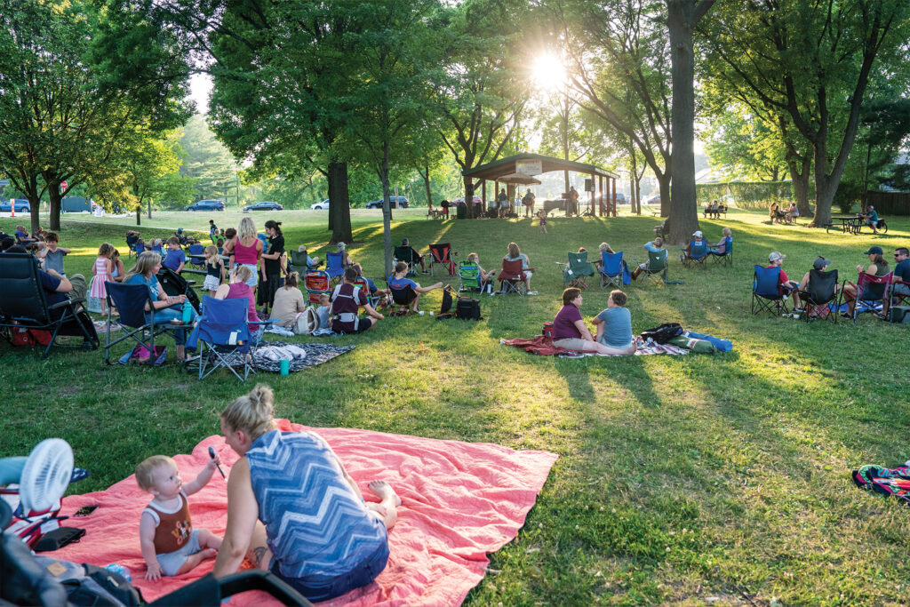 People sitting on chairs and blankets at a park enjoying an outdoor event with a pavilion in the background. Sunlight filters through the trees.