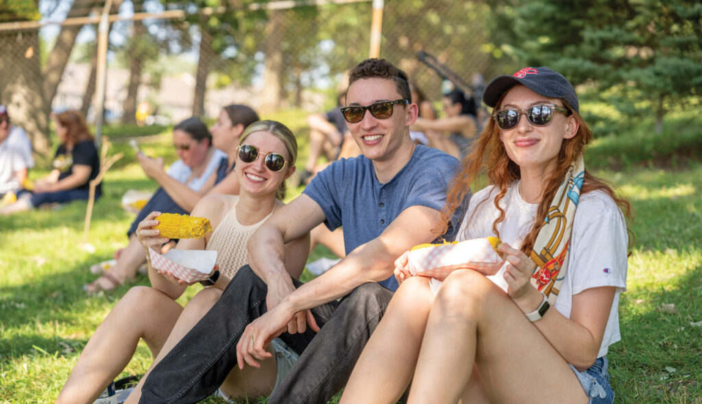 Three people sit on the grass at a park, smiling and enjoying corn on the cob. Other people can be seen in the background.