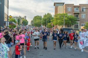 A group of people, including children and adults, walk down a street during a parade, surrounded by spectators. Some participants wear matching T-shirts and some carry banners.