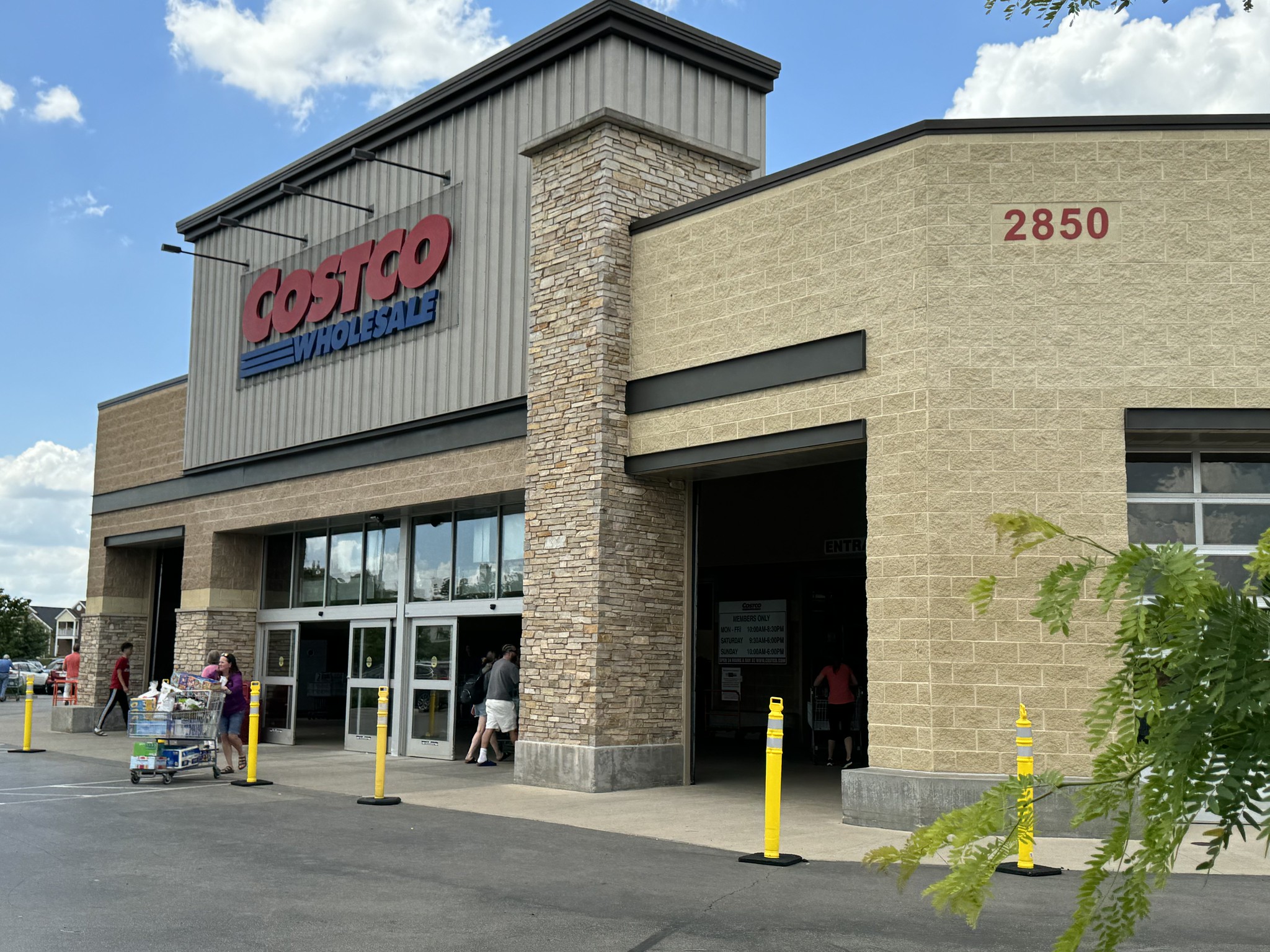 Exterior view of a Costco Wholesale store with shoppers entering and exiting the building. The store's signage and address number, 2850, are visible.