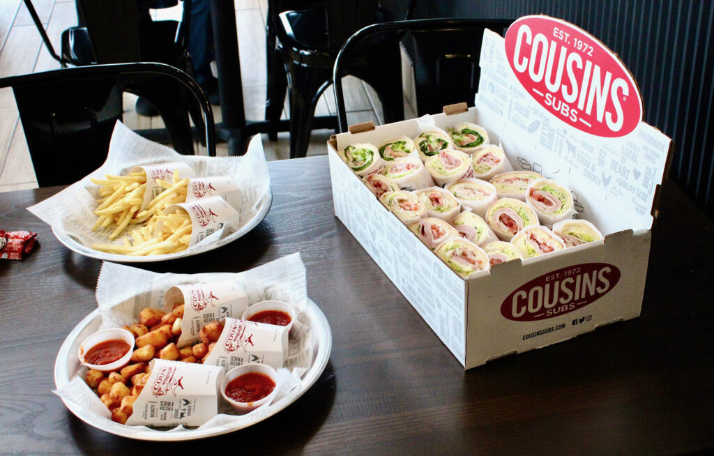 A table with a box of assorted subs from Cousins Subs, a plate of fries, and a plate of fried cheese curds, each accompanied by dipping sauces.