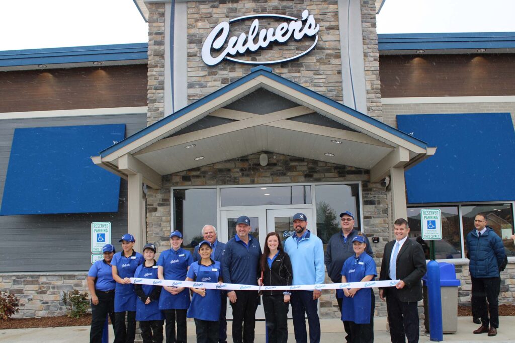 A group of people standing in front of a Culver's restaurant during a ribbon-cutting ceremony. The restaurant sign is visible above the entrance.