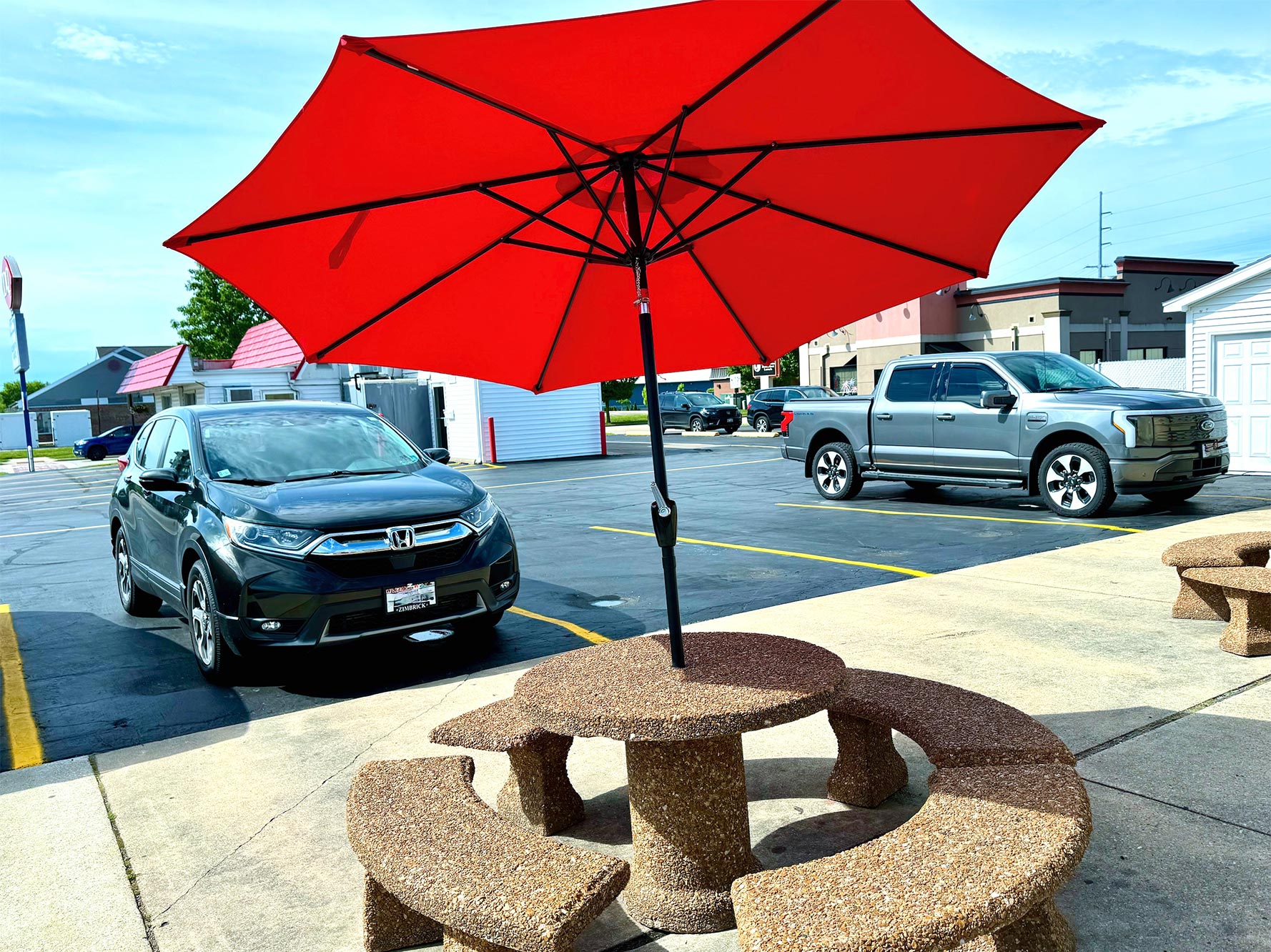 Red umbrella shading a round stone picnic table with benches in a parking lot. A black SUV and a silver truck are parked in the background near some shops.