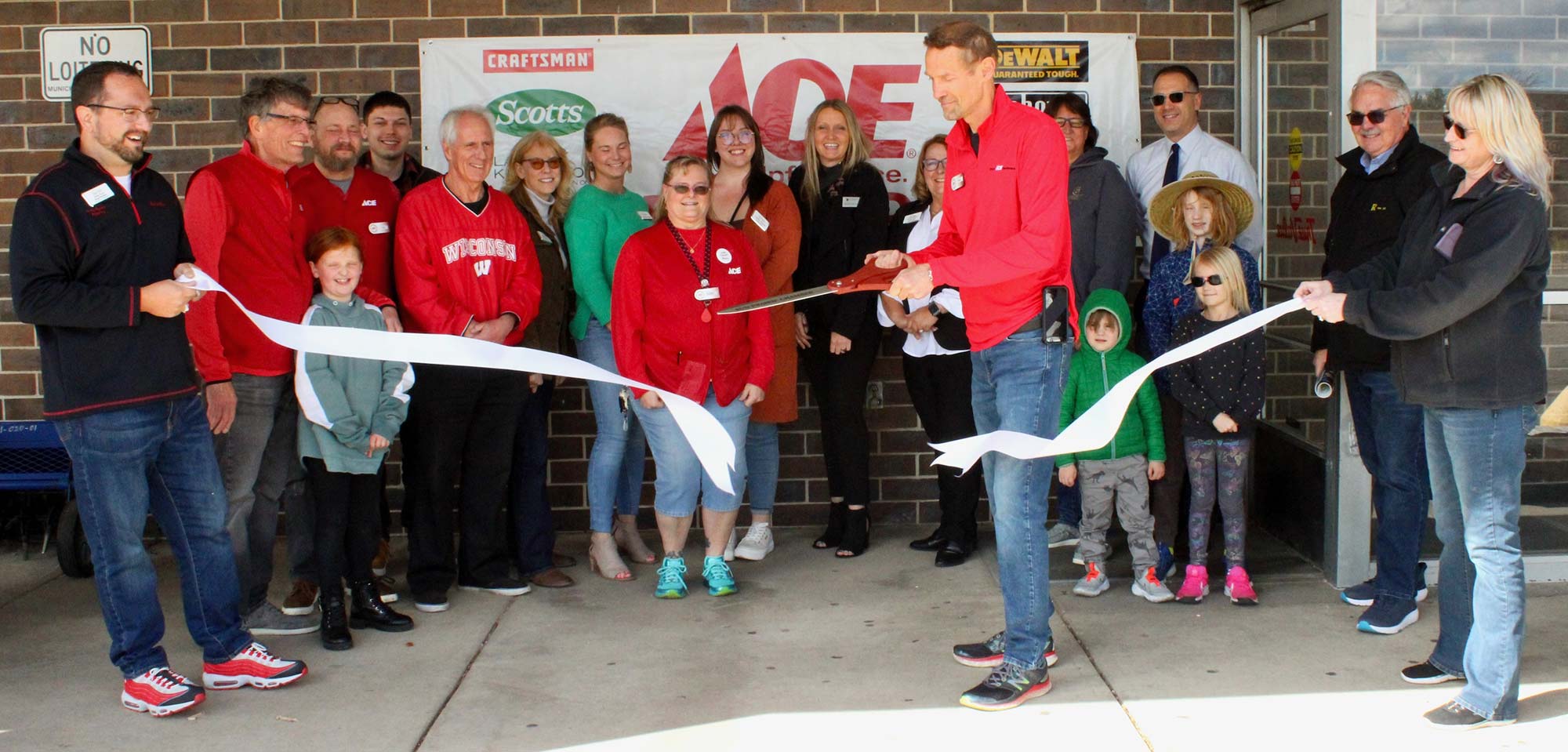 A group of people celebrates a ribbon-cutting ceremony in front of a brick building. A man and woman, both in red shirts, hold large scissors and cut the ribbon, while others watch and take photos.