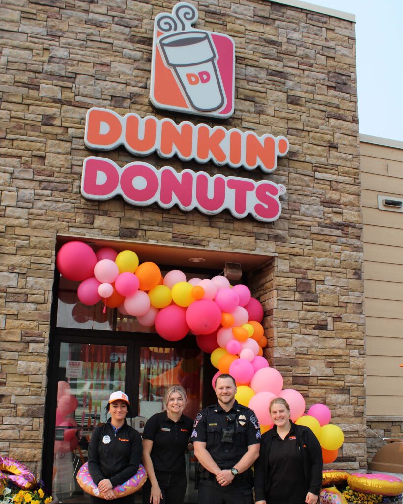 Four people are standing in front of a Dunkin' Donuts entrance decorated with colorful balloons. The group includes three employees dressed in black uniforms and one person in a police uniform.