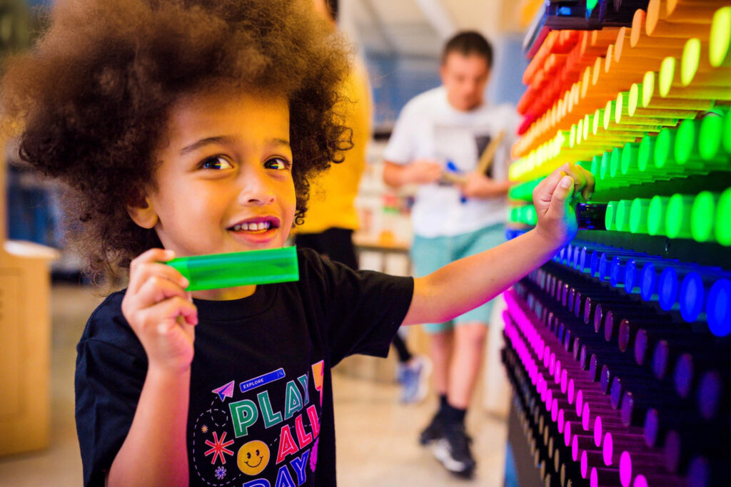 A child smiles while interacting with a colorful wall of illuminated pegs at a play center.