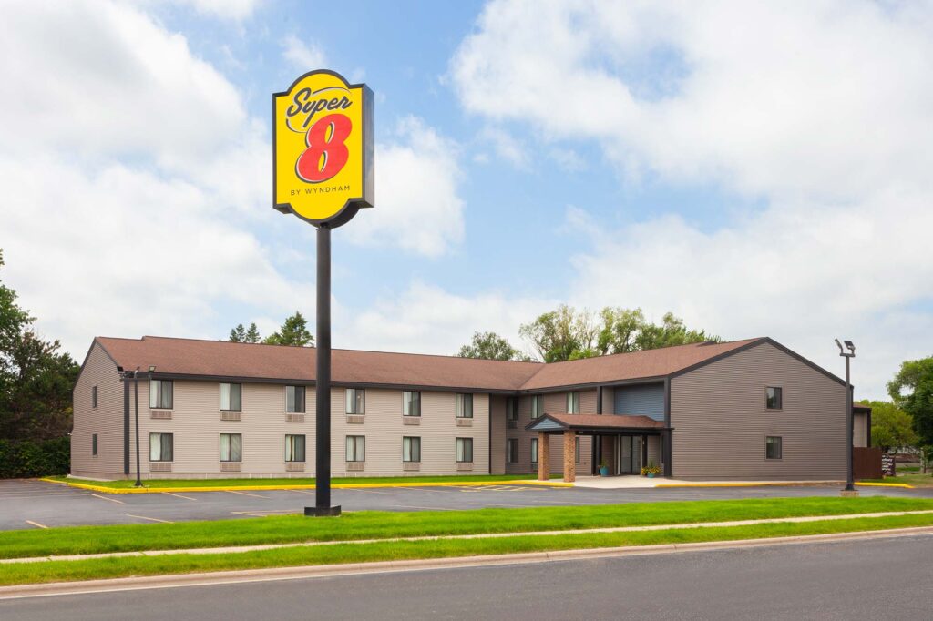 A two-story grey building with a yellow and red sign reading "Super 8 by Wyndham" near a parking lot and bordered by grass.