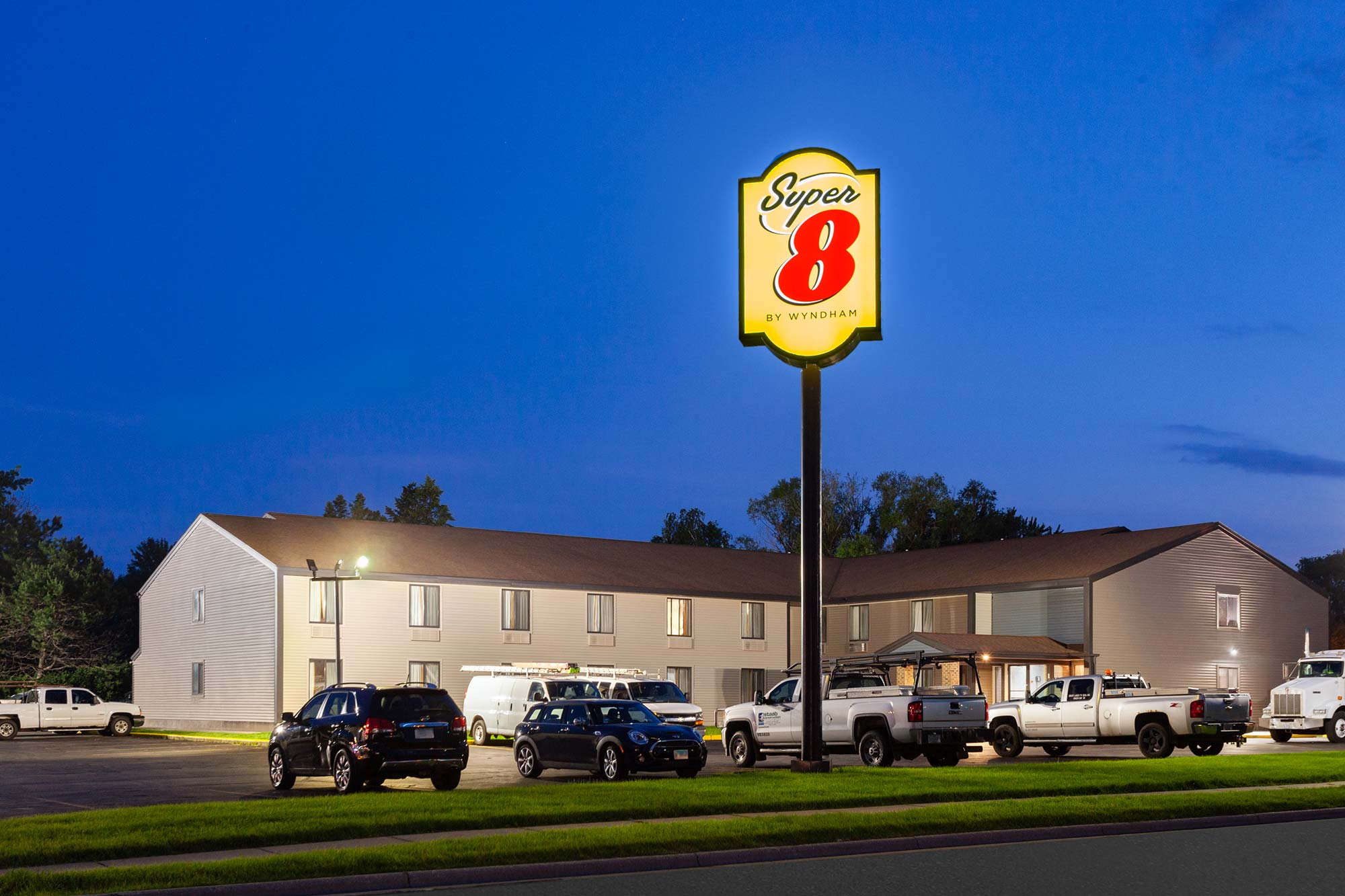 The image shows the exterior of a Super 8 by Wyndham motel at dusk, with several vehicles parked in the lot and the illuminated motel sign prominently displayed.