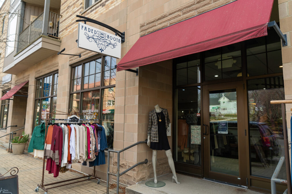 A clothing store named "Faded Roots" with a red awning and a display rack of clothes outside. A mannequin dressed in a black dress stands beside the entrance door.