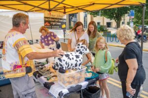 People gathered at a market stall with a cow-themed plush toy and various food items. A "Farmer John’s" sign hangs in the background beneath a yellow canopy.