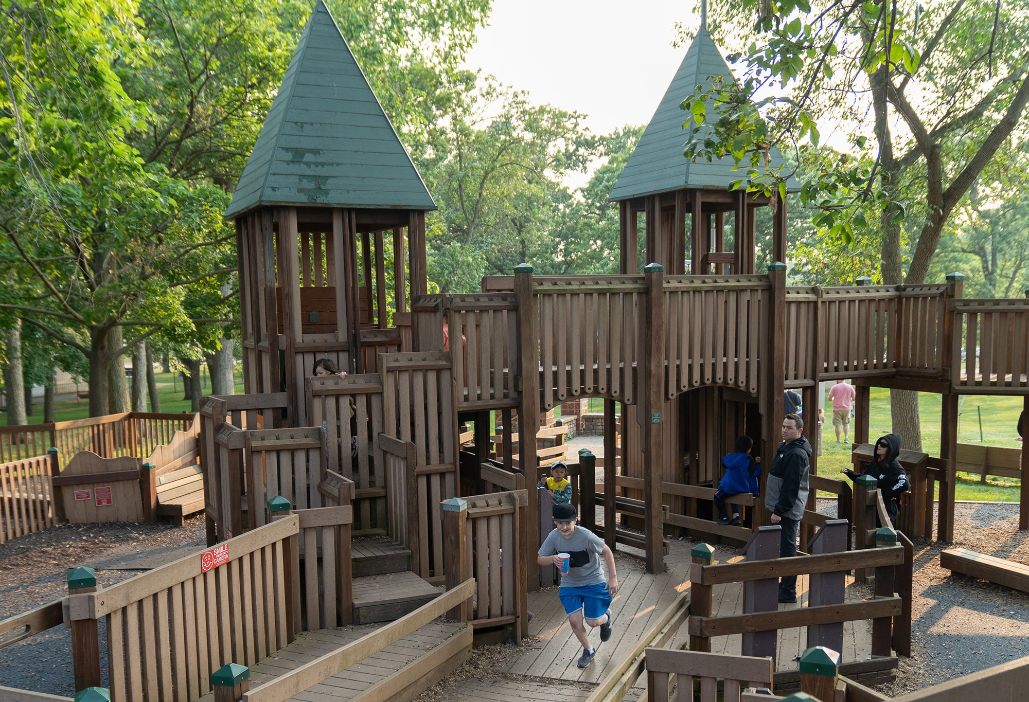 A wooden playground structure with two green-roofed towers is being used by children and supervised by adults. Trees and sunlight are visible in the background.