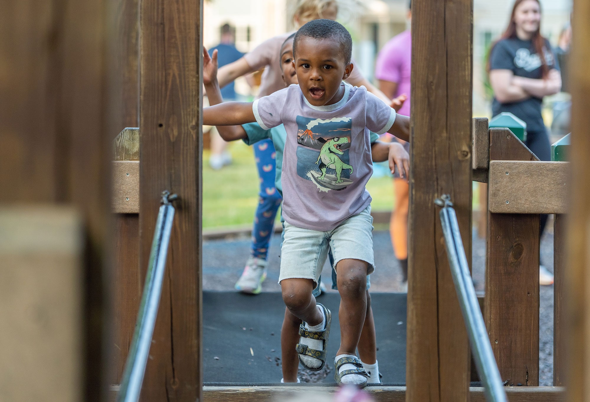 A young boy in a dinosaur T-shirt and sandals runs through a wooden play structure, with other children visible behind him.