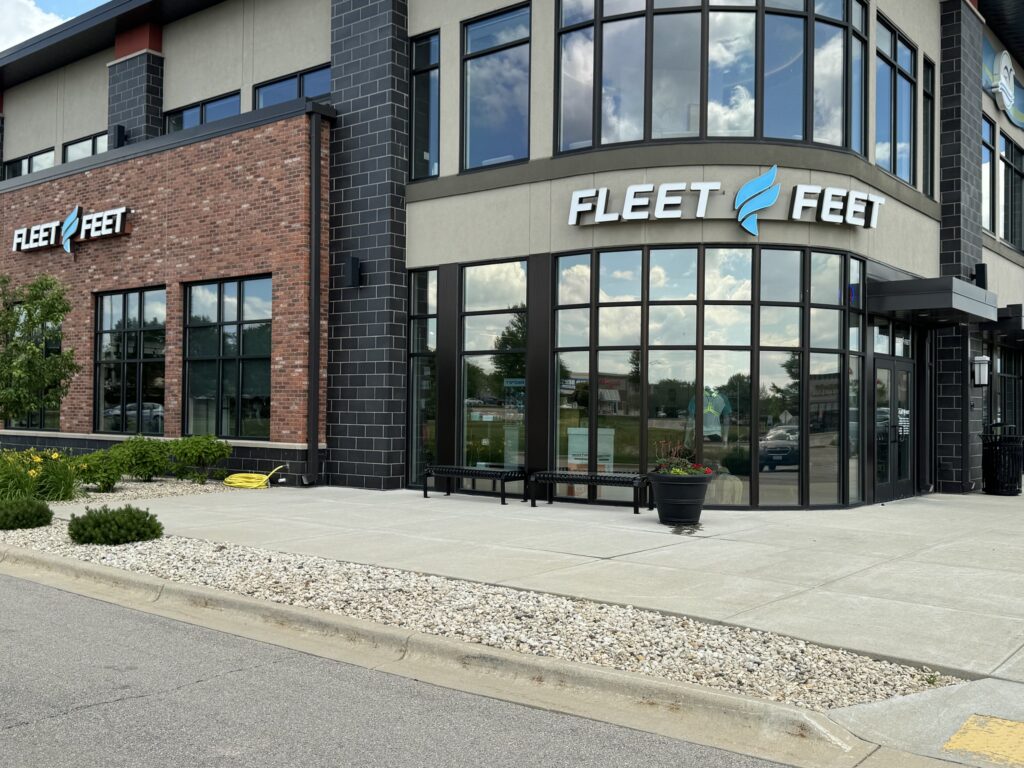 Exterior view of a Fleet Feet store with large windows and signage on a modern brick and stucco building. A sidewalk and landscaped area with shrubs and rocks are in the foreground.