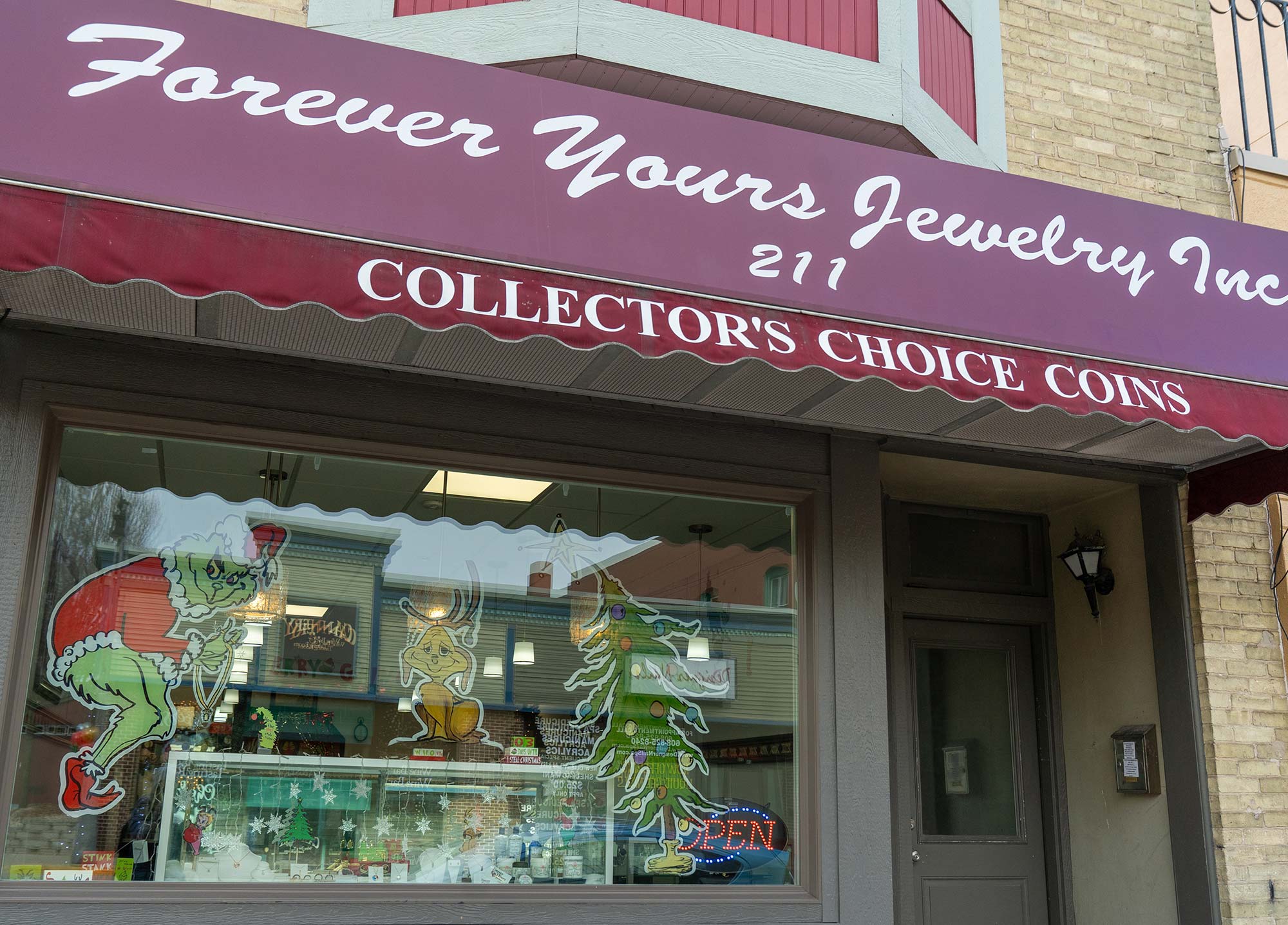 Storefront of "Forever Yours Jewelry Inc" with festive holiday decor, including images of the Grinch and a Christmas tree, and a sign advertising "Collector's Choice Coins." An "OPEN" sign is also visible.