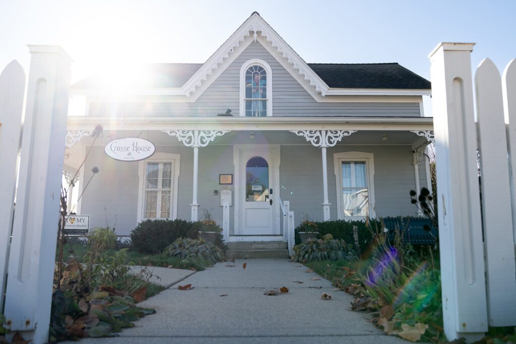 A quaint grey house with white trim, featuring a steep roof, decorative gables, a small porch, and a gated white picket fence, bathed in sunlight.