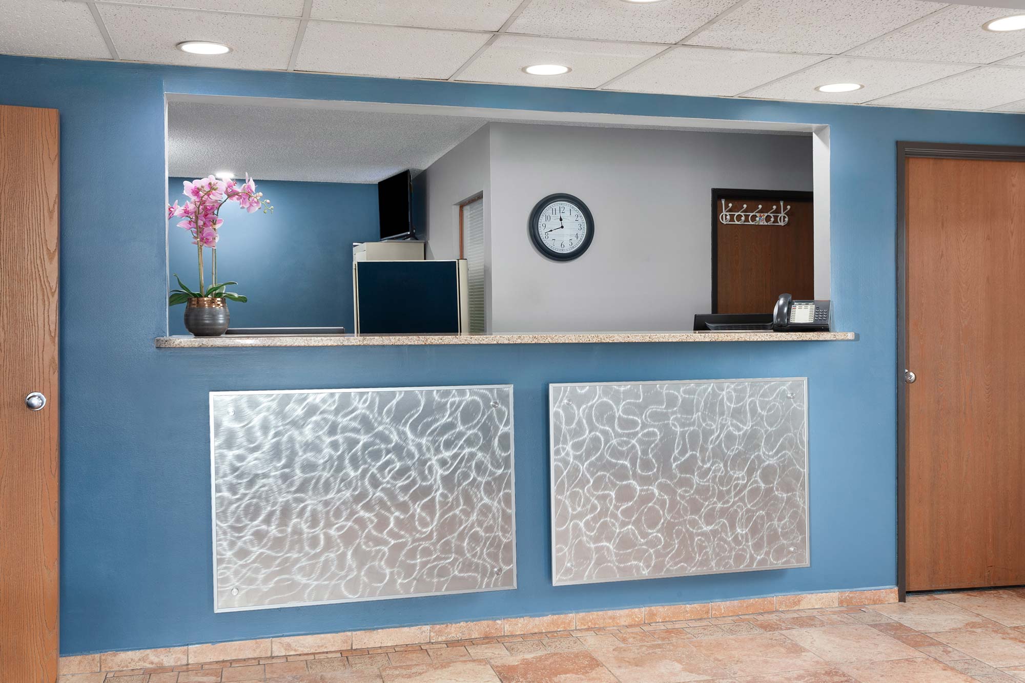 A reception desk in an office lobby with a clock, phone, and a potted orchid. The desk has a granite counter and metallic panels. There are two wooden doors on either side.