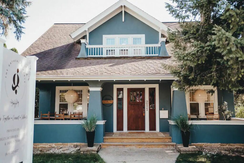 A two-story blue house with a wooden front door, porch, and warm white trim. The yard is landscaped with stone accents and greenery. There is a sign on the left side of the image.