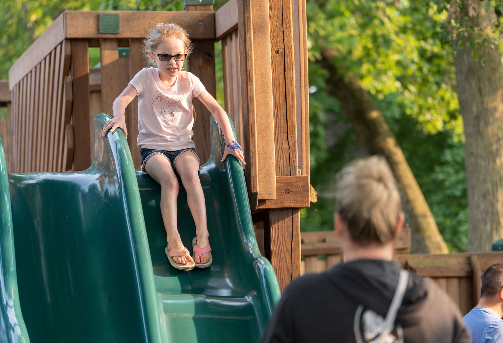 A young girl wearing glasses and a pink shirt slides down a green playground slide while an adult watches from below.