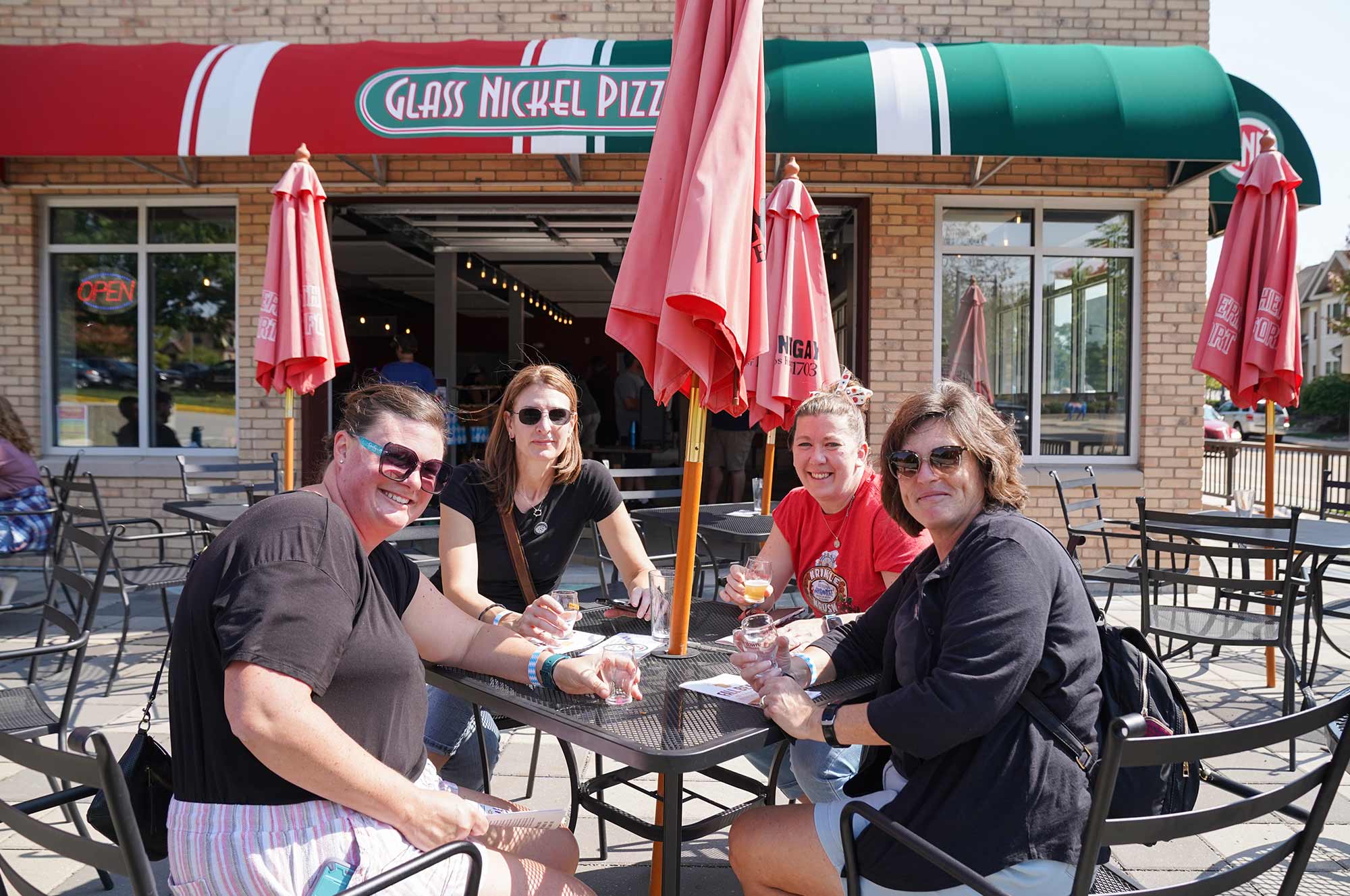 Four people sitting at an outdoor table with drinks in front of a restaurant called "Glass Nickel Pizza Co." on a sunny day.