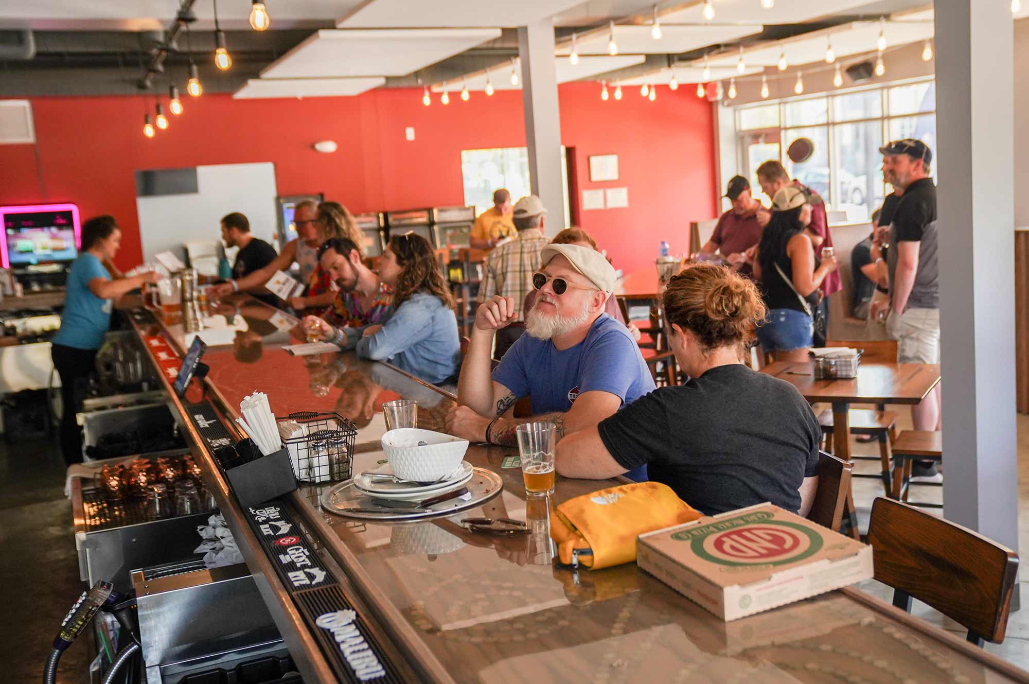 People are sitting and standing at a bar and restaurant, engaging in various activities. The ambiance is casual with warm lighting and a mix of modern and rustic decor.