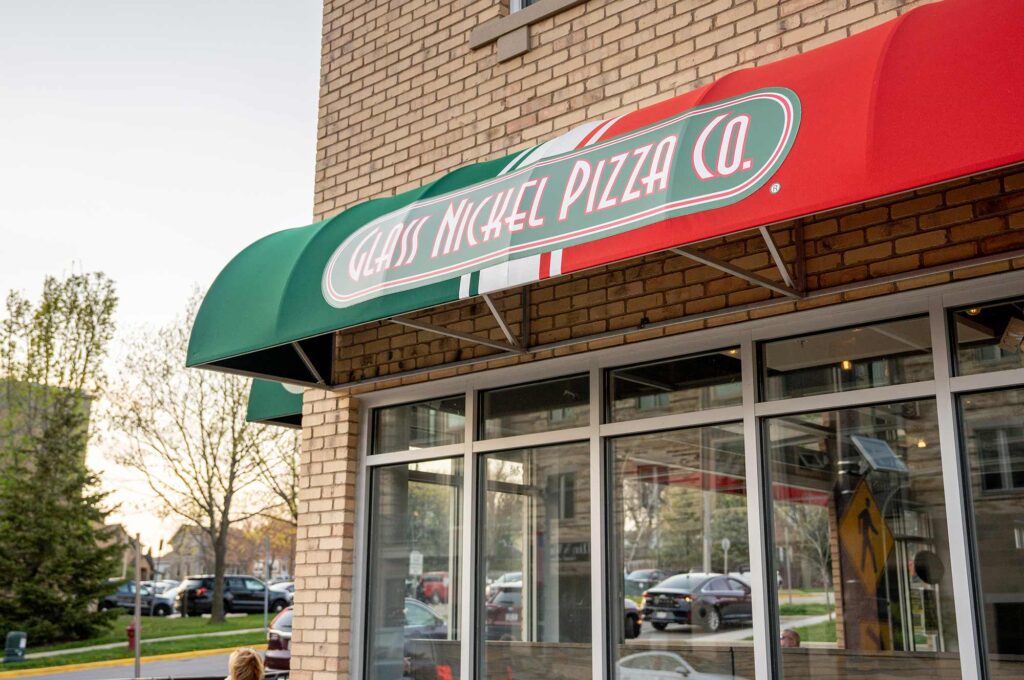 Exterior of a pizza restaurant with a sign reading "Glass Nickel Pizza Co." The storefront has a red, white, and green awning and large glass windows. Trees and parked cars are visible in the background.