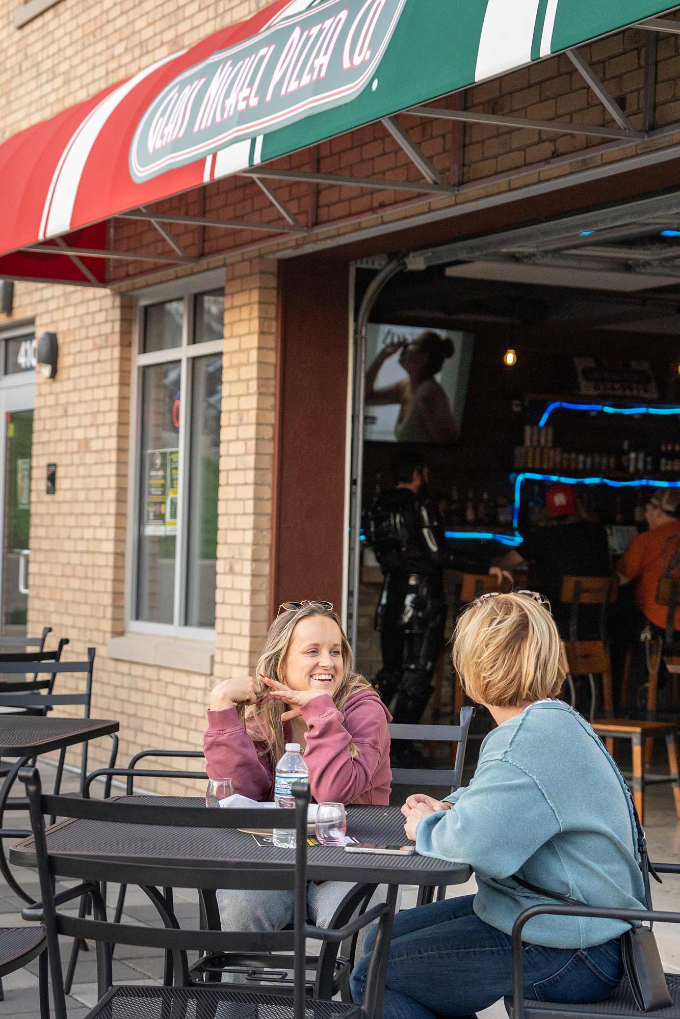 Two women sit at an outdoor table, talking and smiling, outside of Great Wheel Pizza Co. The establishment's awning is red, white, and green. A person is seen inside the establishment near the bar.