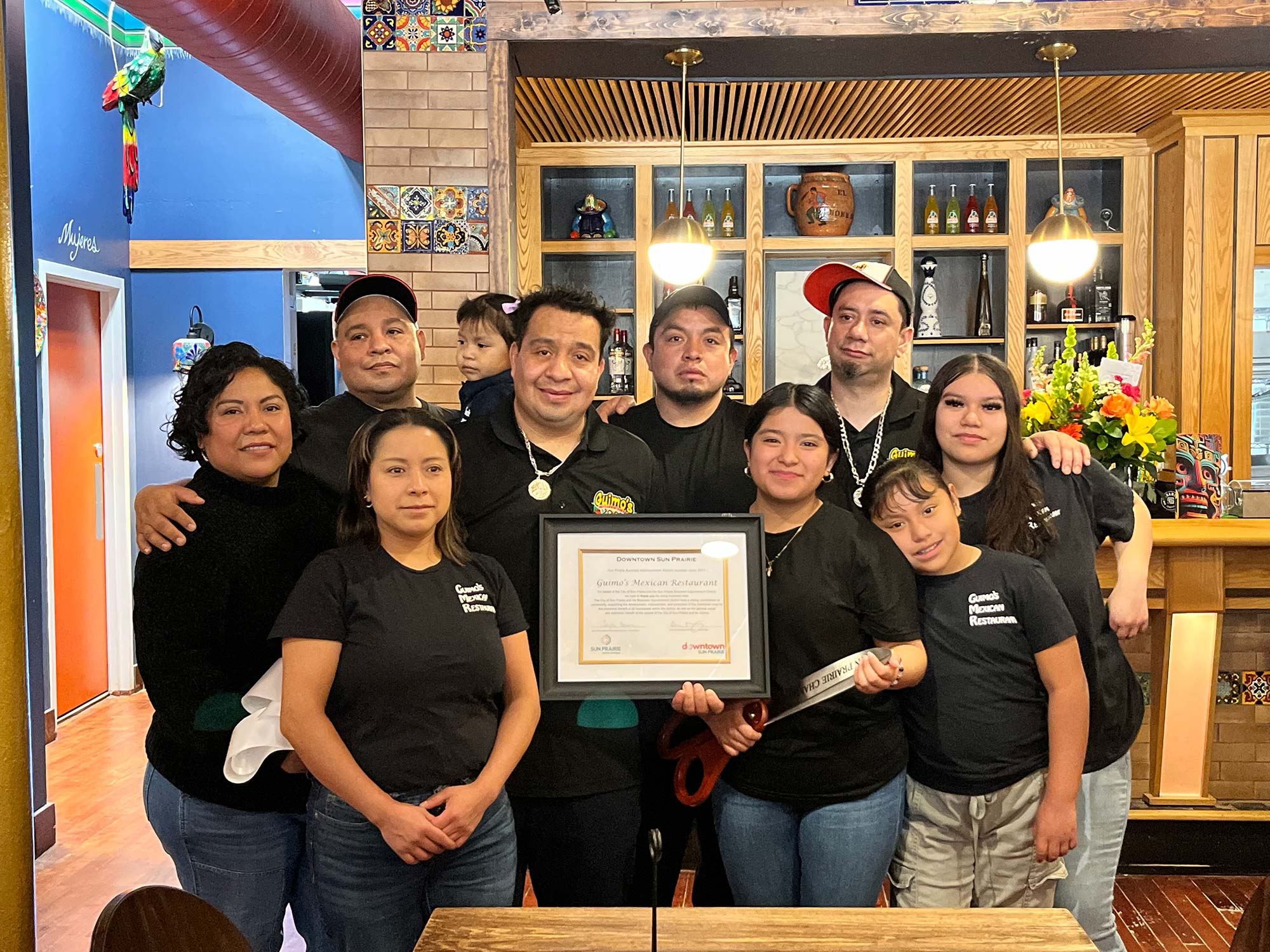 A group of people wearing black shirts stand together inside a restaurant, holding a framed certificate. Some are smiling, and the background shows a bar area and decorative tiles.