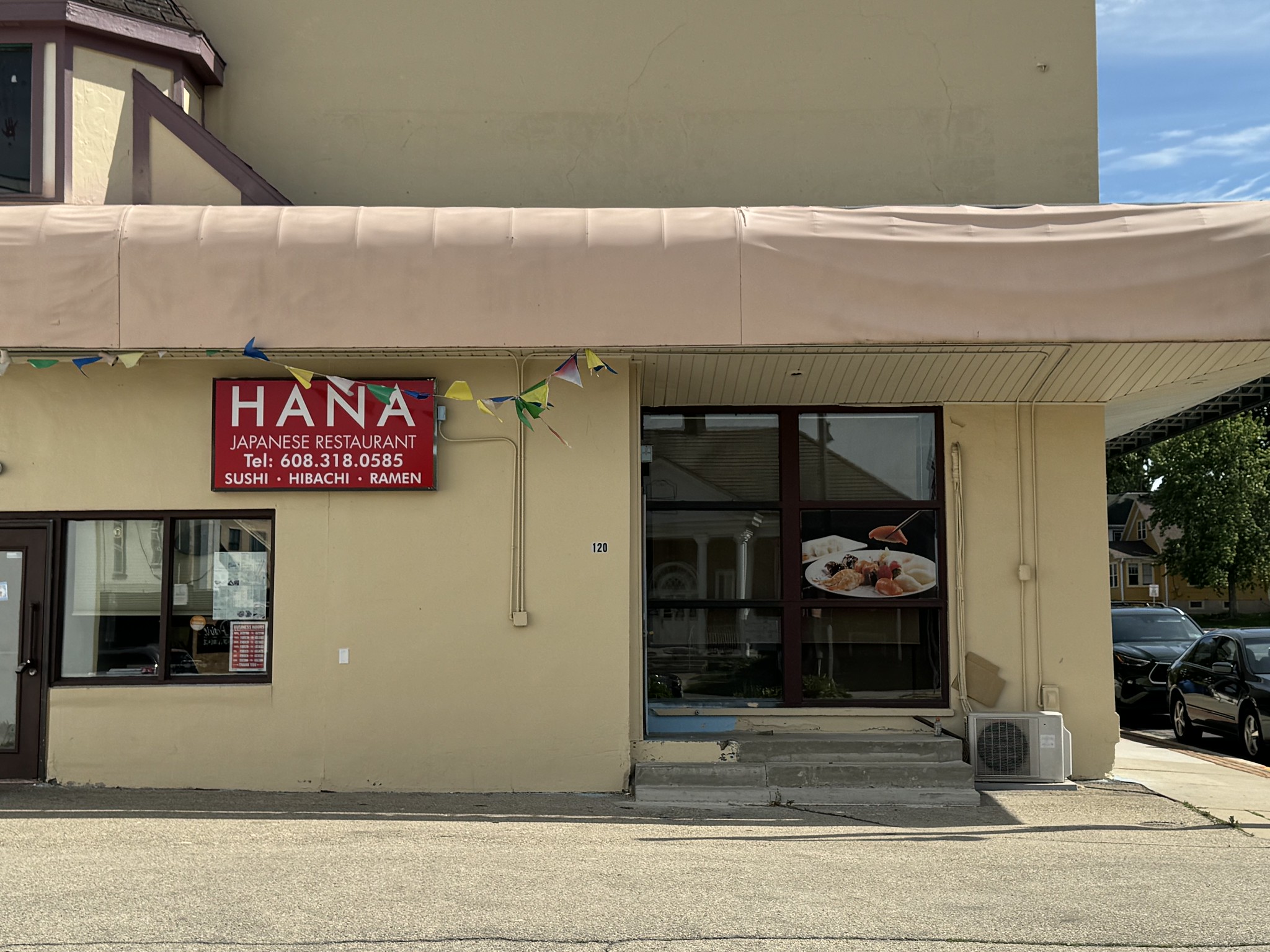 Exterior of Hana Japanese Restaurant with a sign listing sushi, hibachi, and ramen. An image of food is displayed on a large window next to the entrance steps. Bunting decorates the awning above.