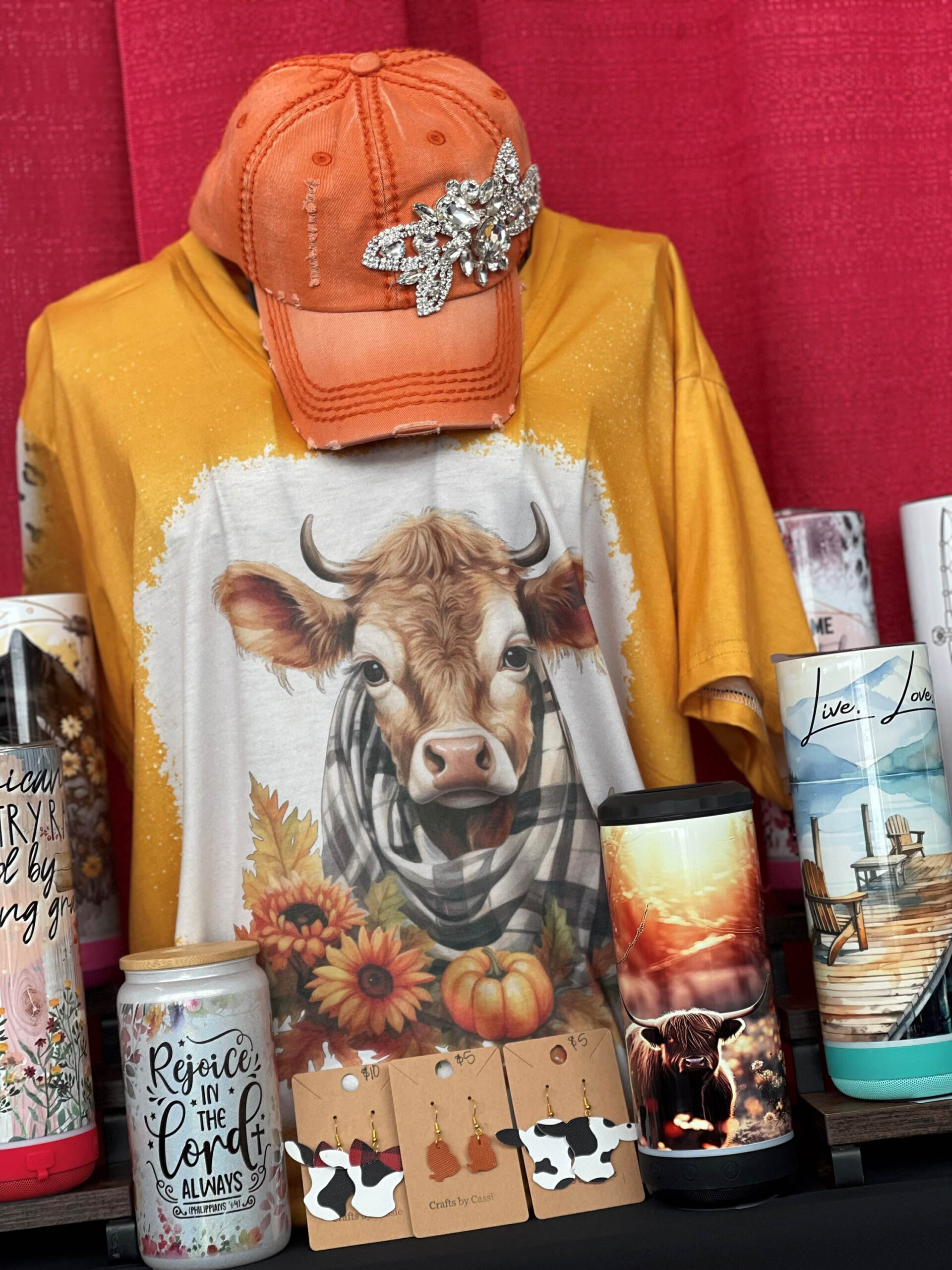 Display featuring a yellow t-shirt with a cow print, an orange cap adorned with a brooch, various tumblers, earrings, and a candle, all set against a red fabric backdrop.