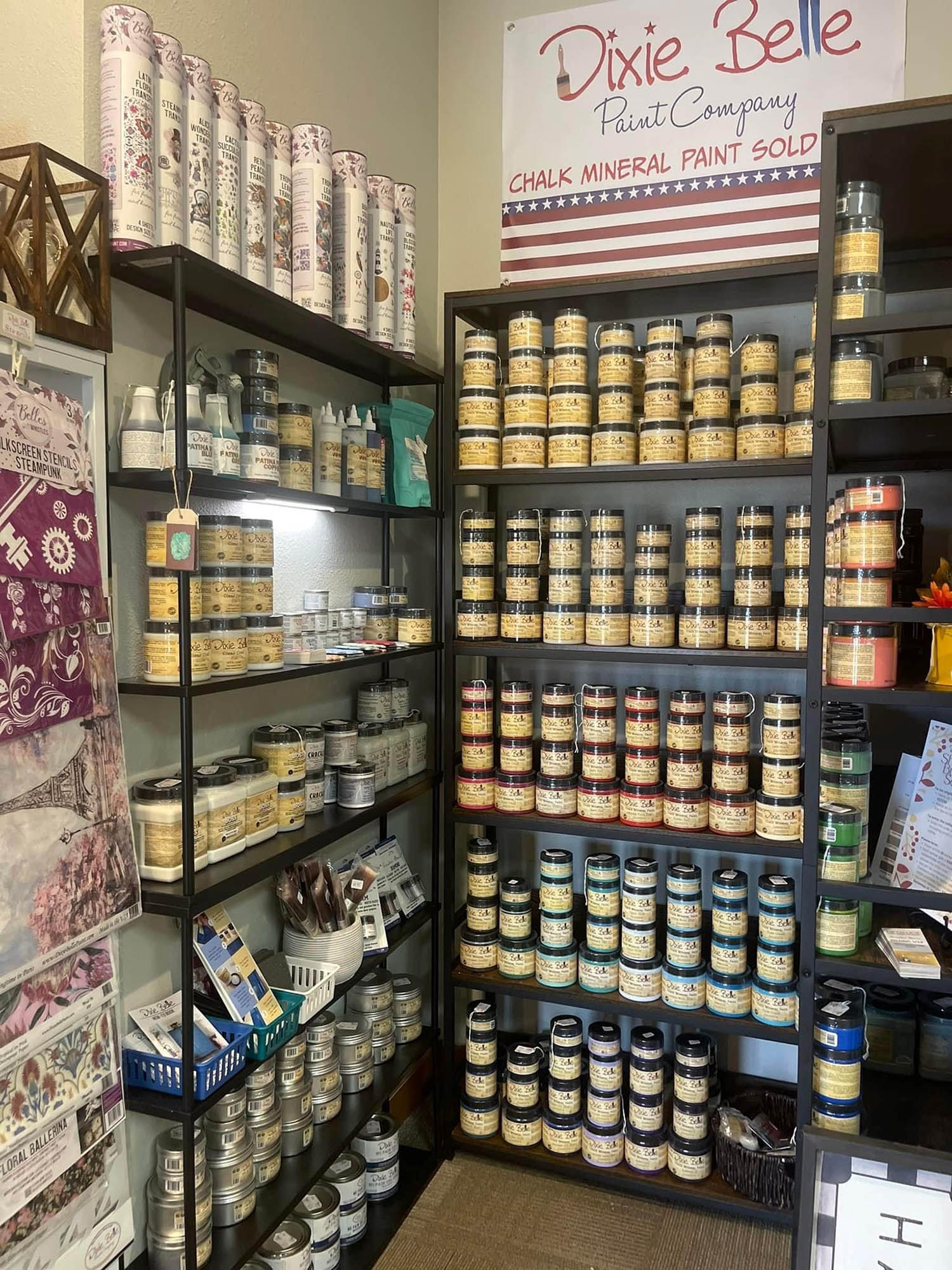 Display of Dixie Belle chalk mineral paints on black shelves in a store. Various colors and sizes of paint containers are neatly organized. A banner above the shelves reads "Dixie Belle Paint Company.