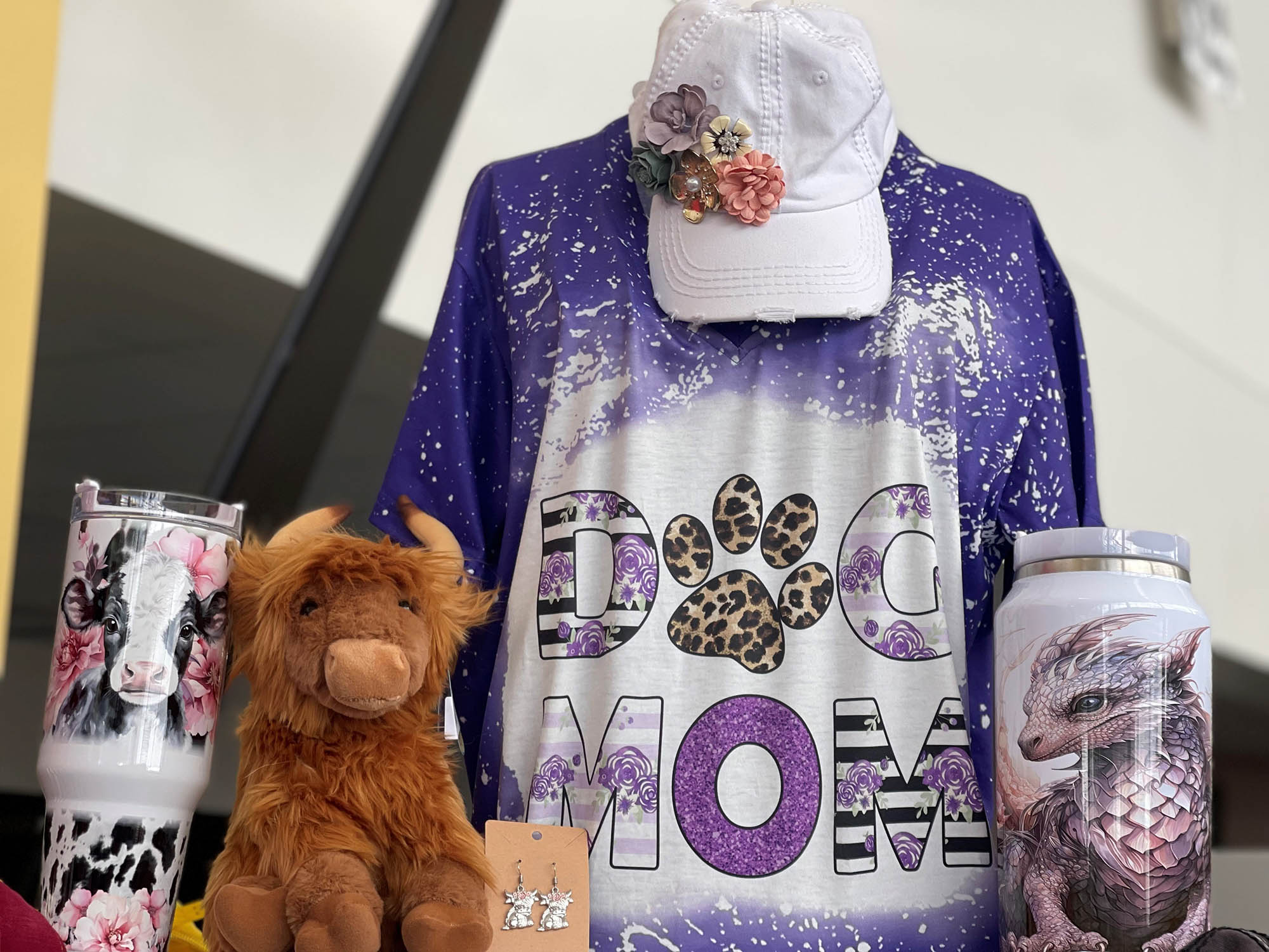 Display of items including a purple "Dog Mom" shirt, hat with floral adornments, cow and dragon-themed cups, small plush toy, and earrings.