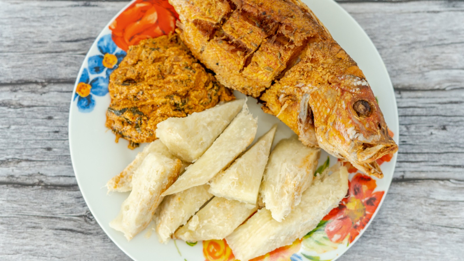 A plate with grilled fish, a side of orange-colored paste, and several pieces of sliced boiled cassava on a floral-patterned dish.